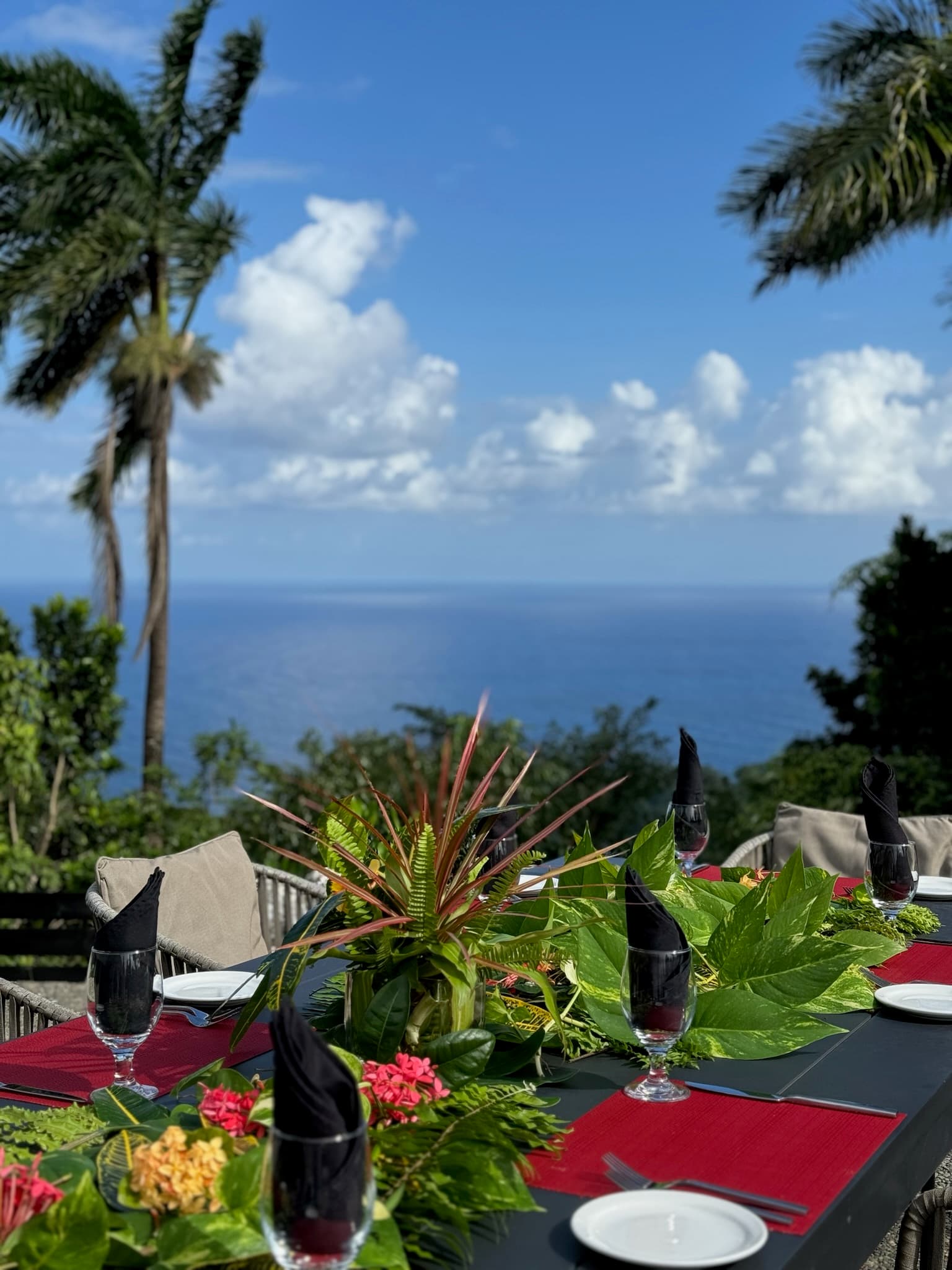 An outdoor dining table set with black napkins, red placemats, tropical floral arrangements, and a sweeping ocean view framed by palm trees and blue sky.