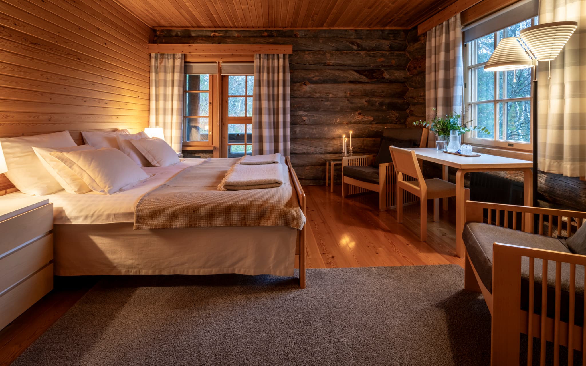 A cozy rustic bedroom with a large bed, wooden walls and ceiling, log wall details, and a small seating and desk area by the window.