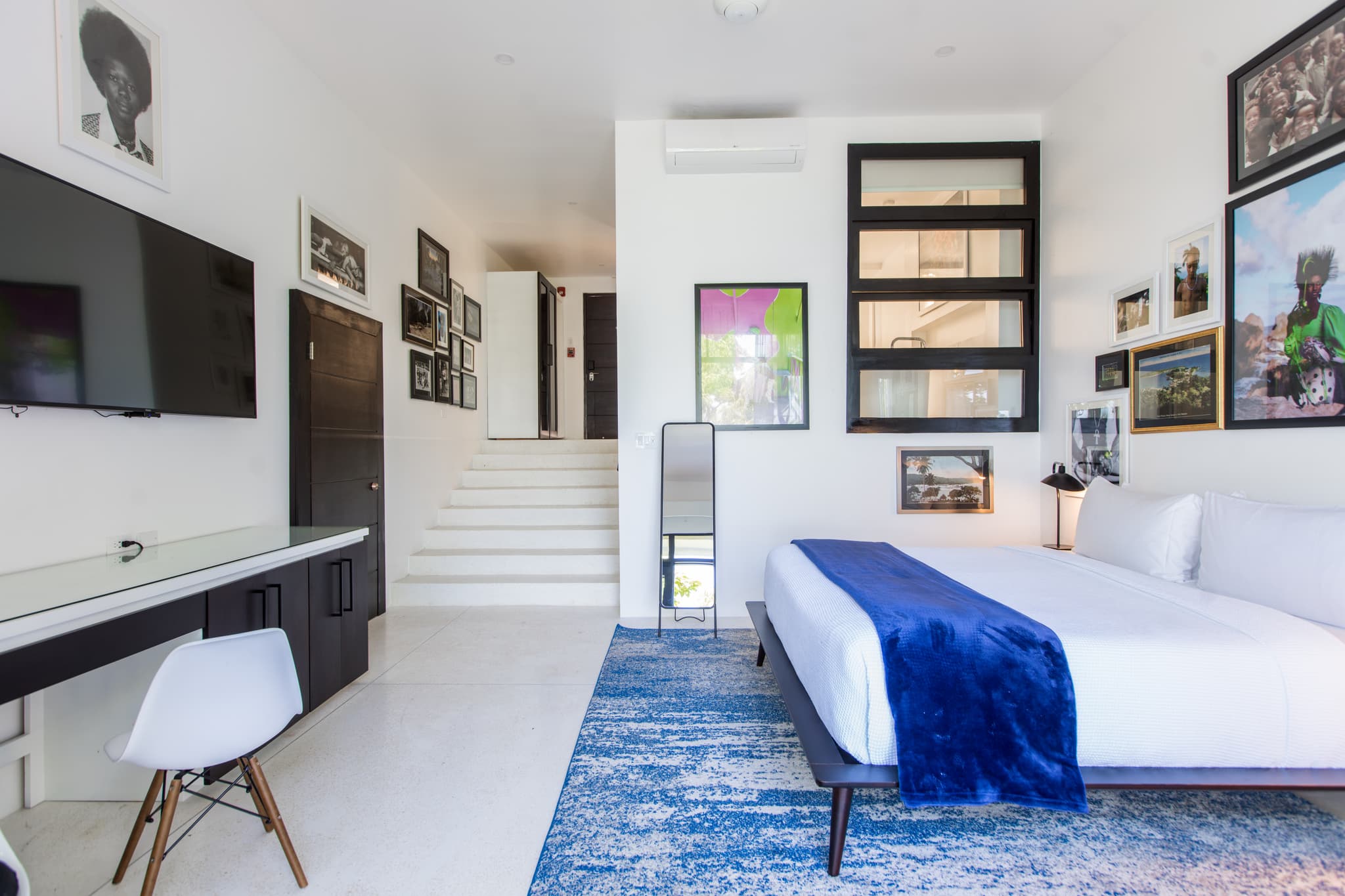 A bright modern bedroom features a large bed with white linens and a blue throw, framed wall art, a desk area, and a staircase in the background.