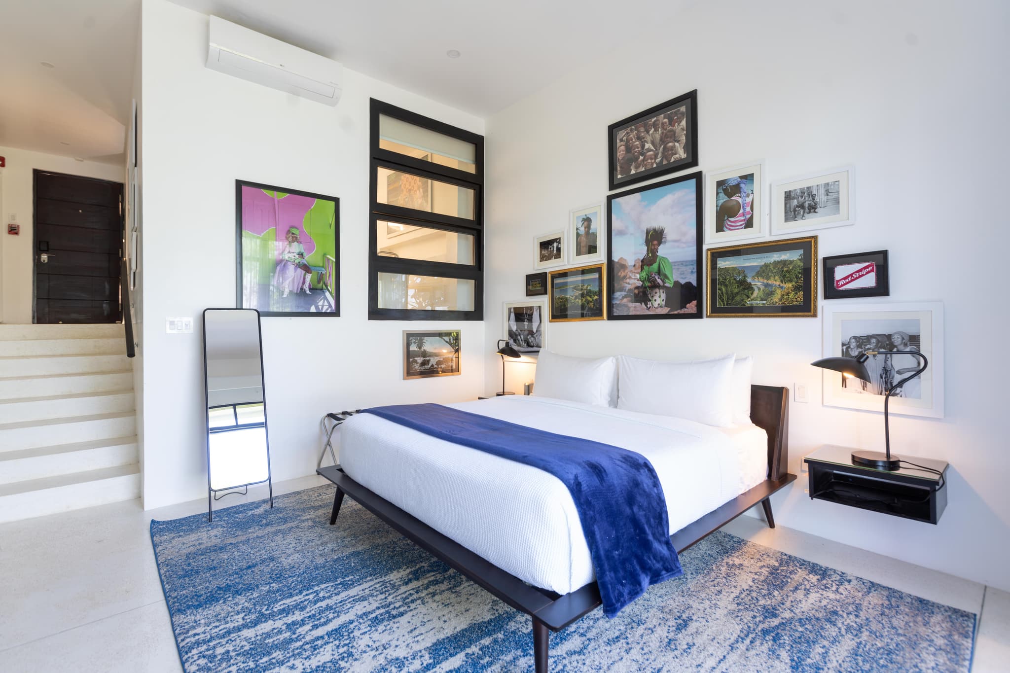 A modern bedroom with a low platform bed, blue throw, patterned rug, wall art, and a mirror beside a staircase.