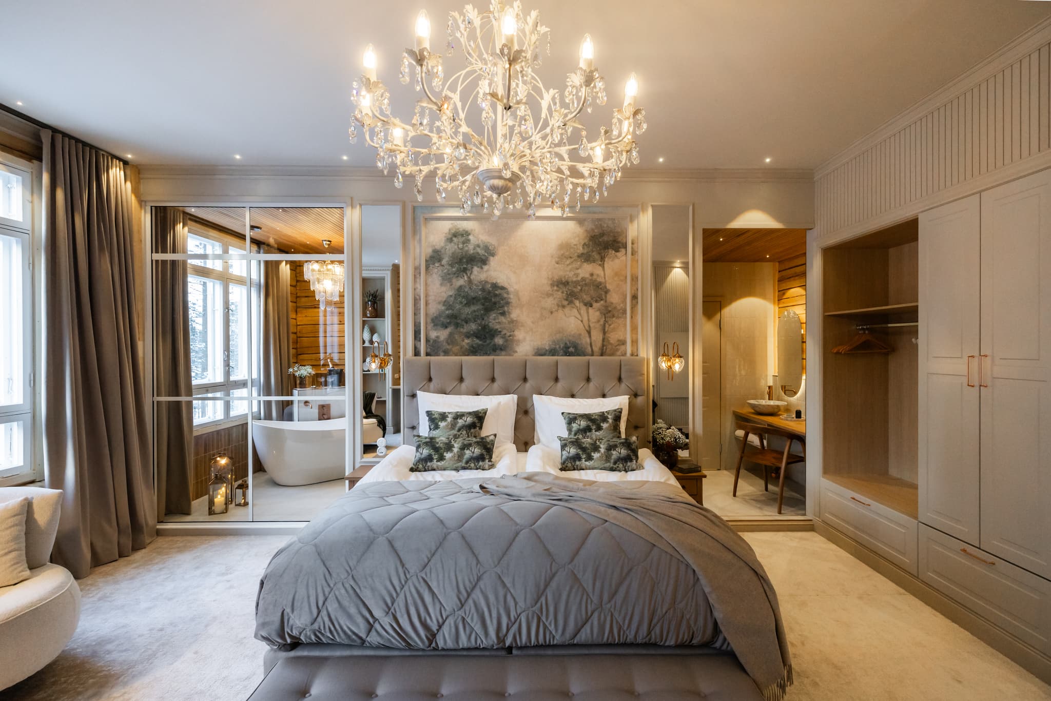 A luxurious bedroom with a tufted bed, crystal chandelier, mirrored wall, and adjacent bathroom and dressing area.