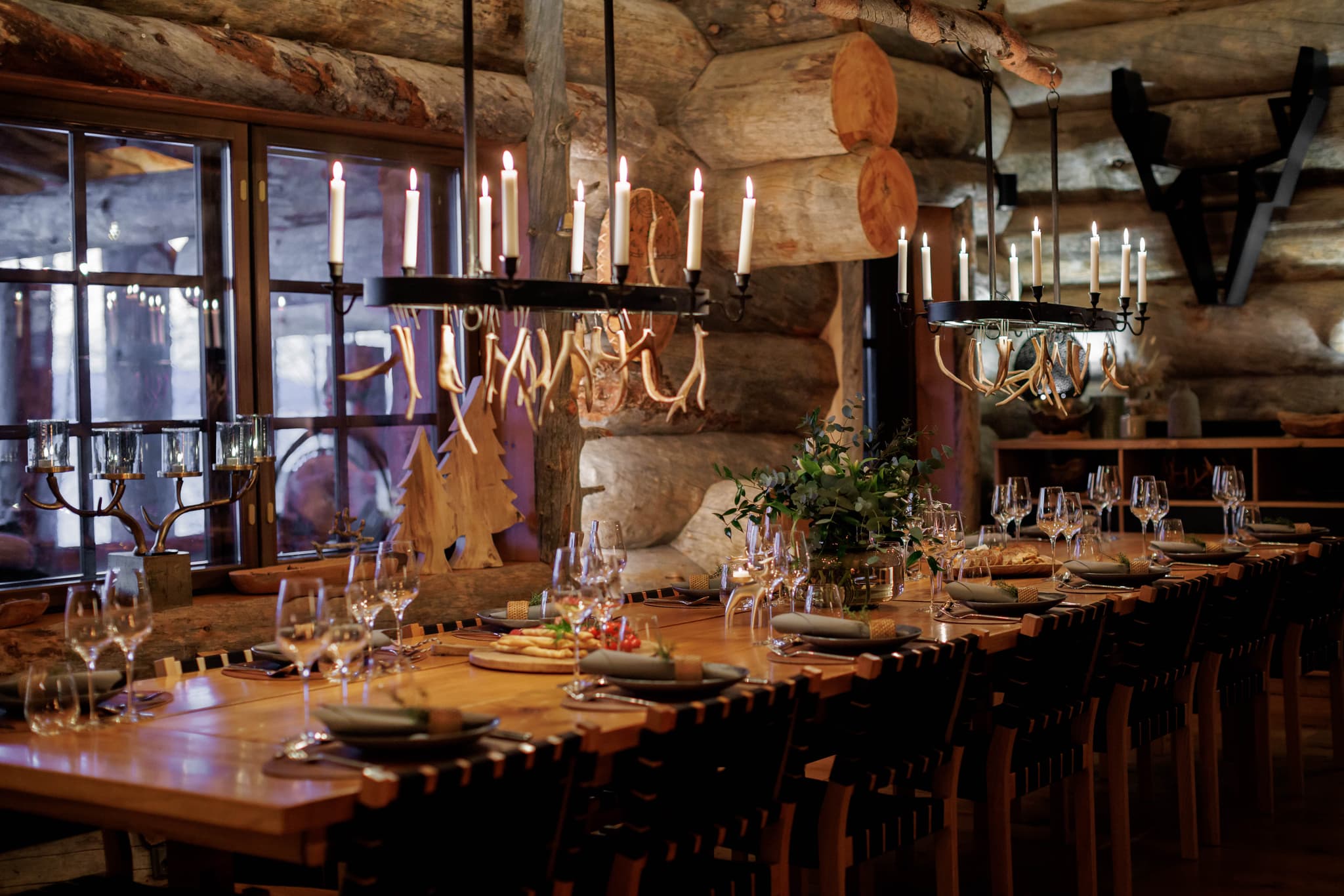 A warmly lit rustic dining room features a long set table with candles, glassware, and greenery beneath log walls and hanging chandeliers.