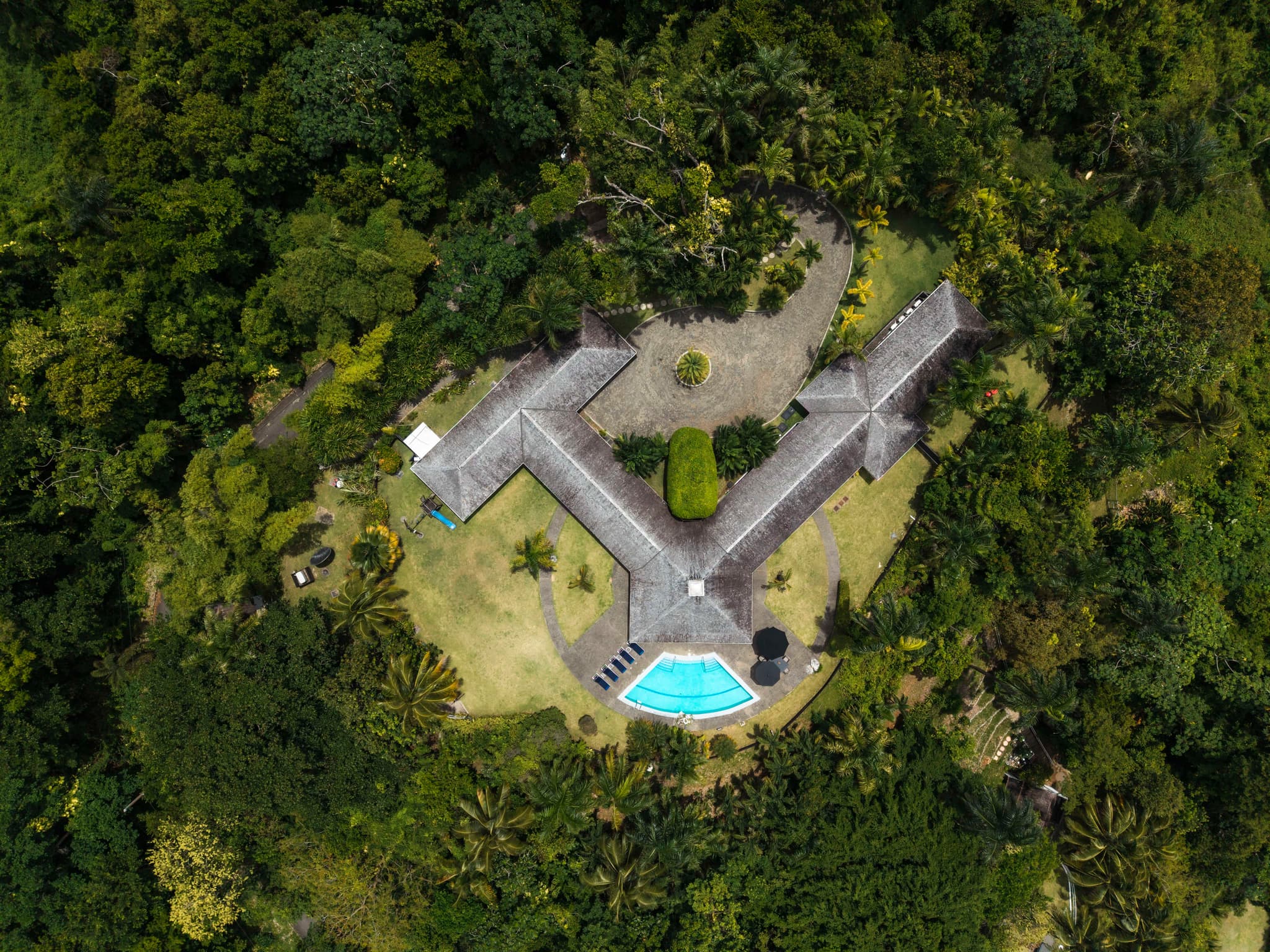 An aerial view shows a large villa with a curved swimming pool surrounded by dense tropical forest and landscaped grounds.