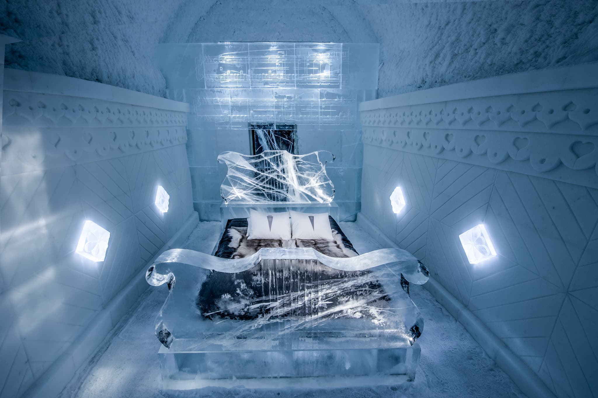An ice bedroom features a translucent ice bed with fur bedding, carved wall details, and glowing geometric lights in a blue-lit frozen chamber.