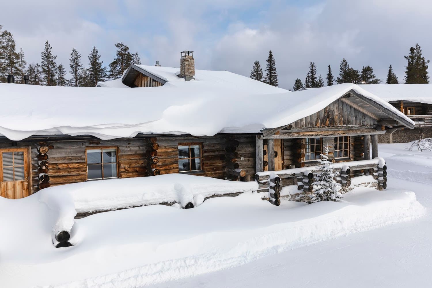 A snow-covered log cabin sits in a deep winter landscape beneath a cloudy sky.