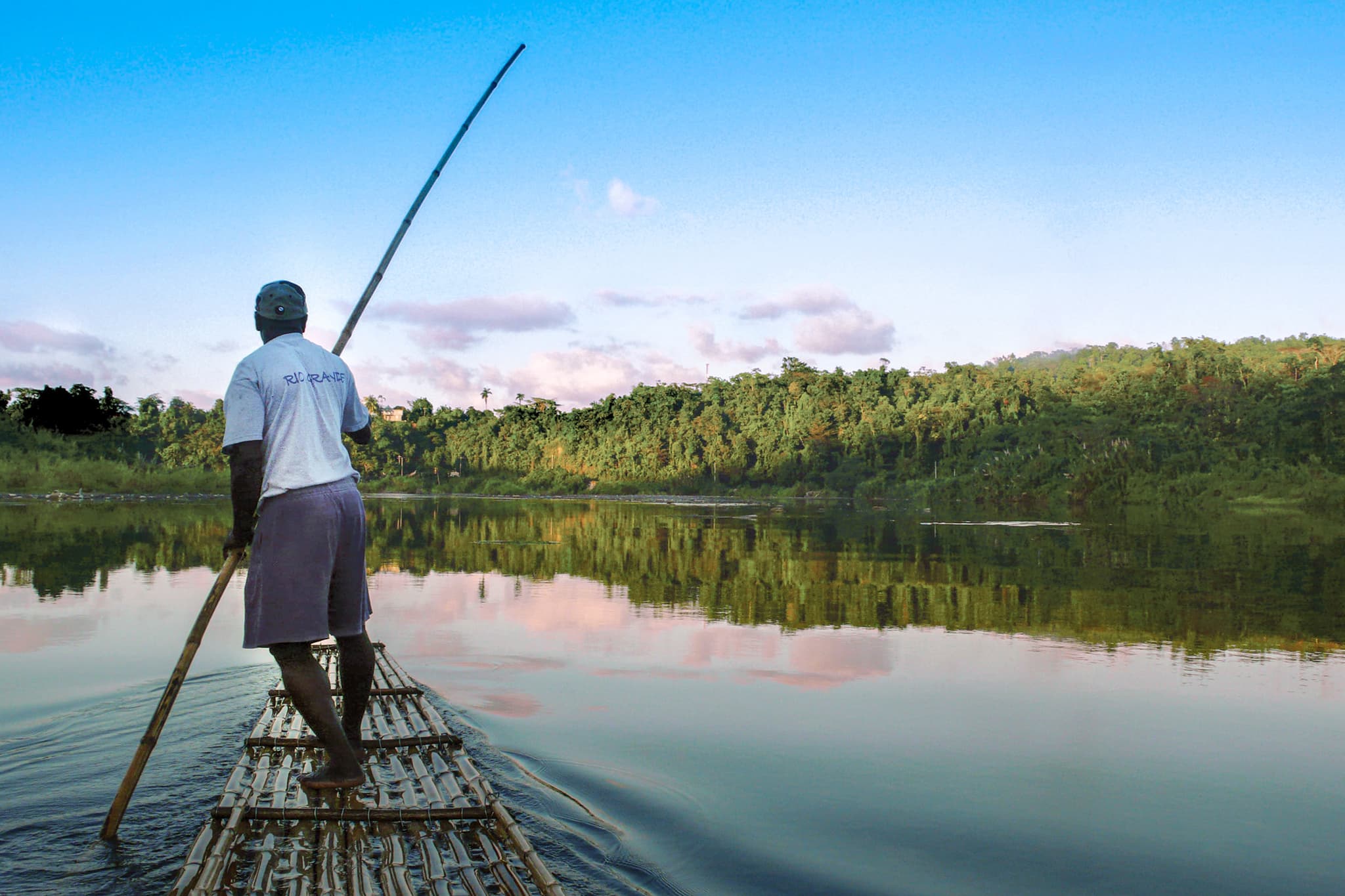 A man stands on a bamboo raft and uses a long pole to steer across a calm river with forested banks and a reflective sky.
