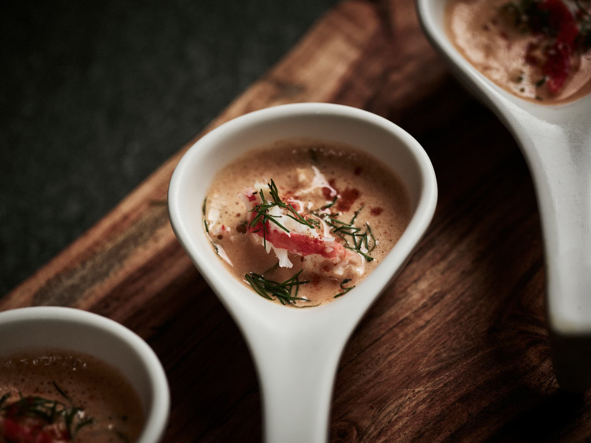 A close-up of a plated seafood soup served in white spoon-shaped bowls on a wooden board.