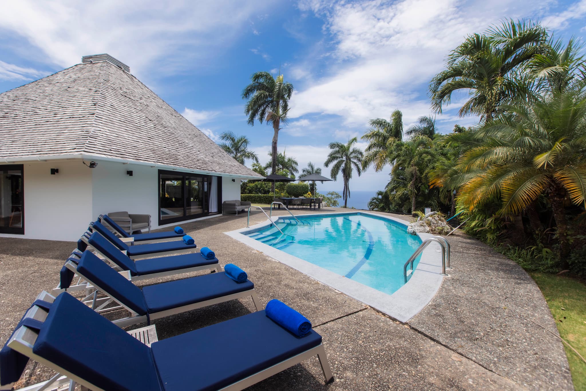 A tropical villa terrace features a swimming pool, blue sun loungers, palm trees, and a distant ocean view under a bright sky.
