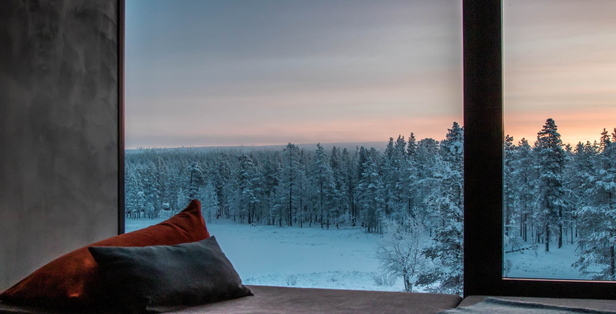 A cozy interior window seat overlooks a snowy forest at sunset.