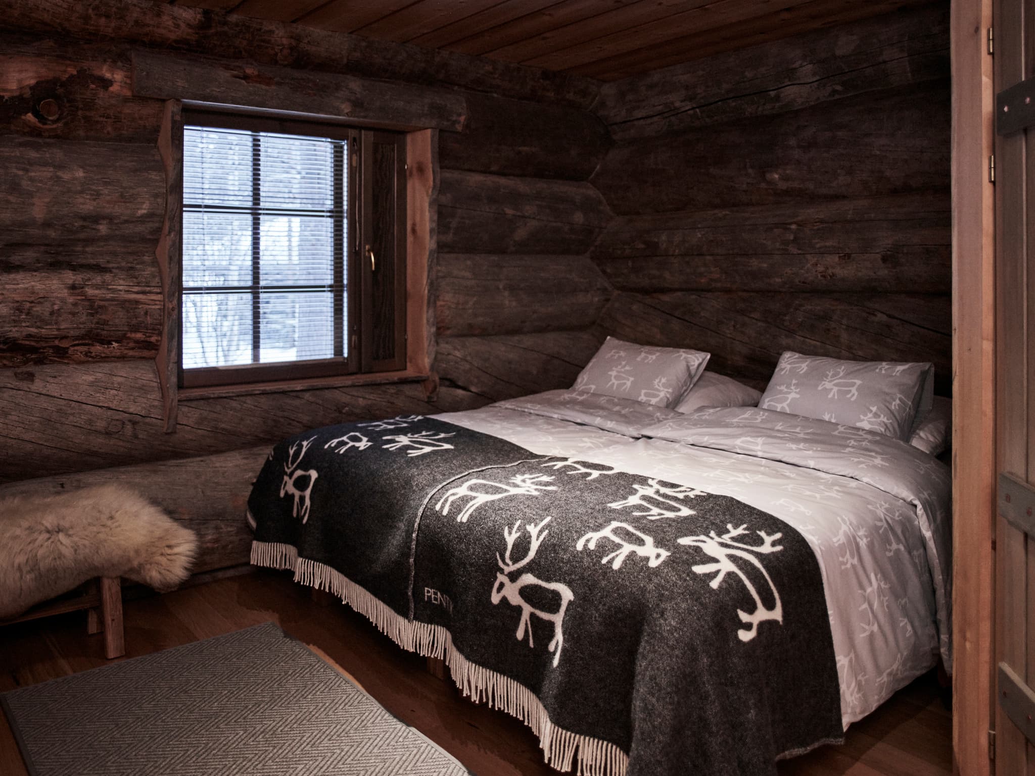 A cozy log cabin bedroom features a neatly made bed with patterned bedding, a small window with blinds, and rustic wood walls and ceiling.