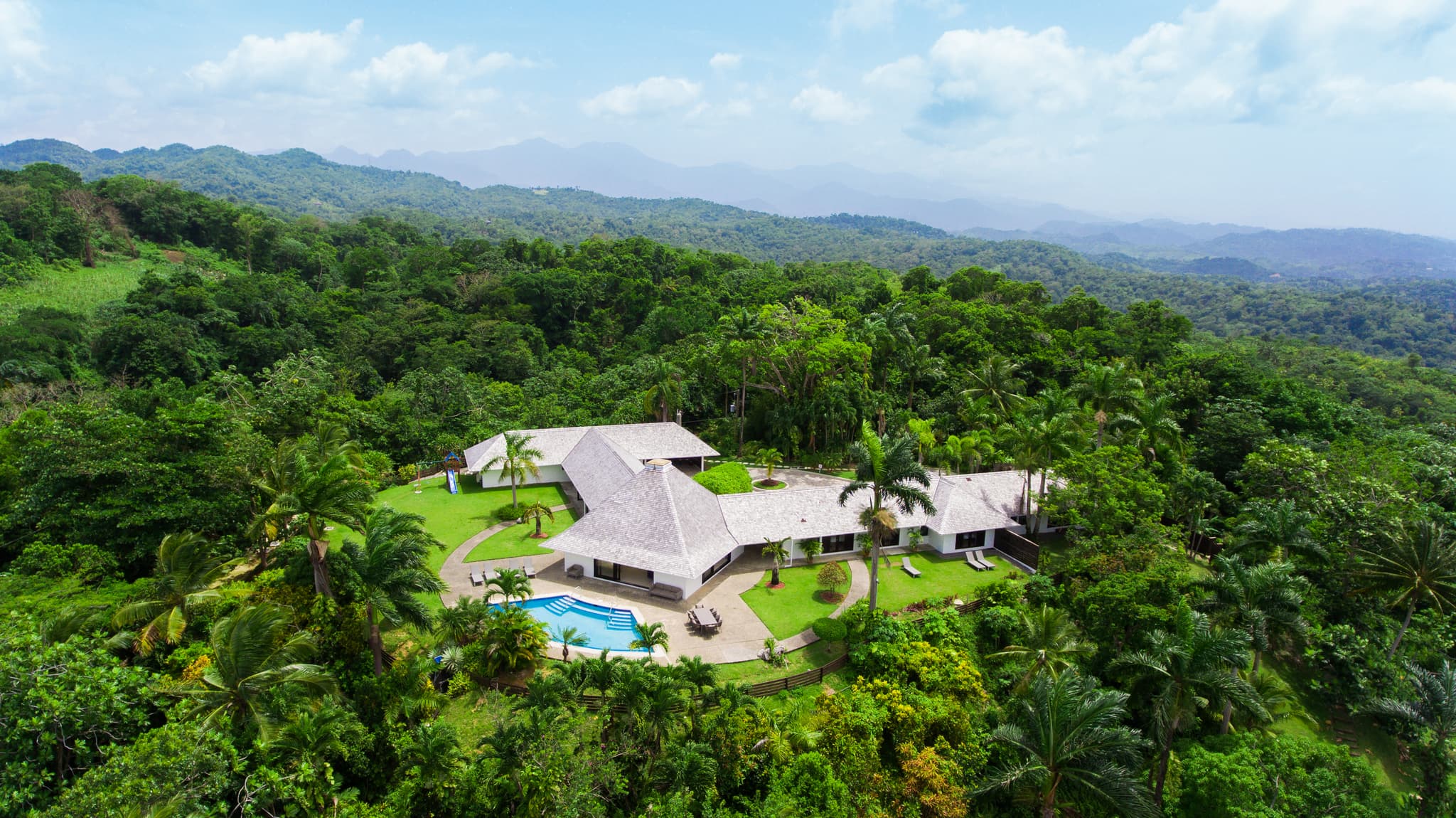 An aerial view shows a white-roofed villa with a swimming pool surrounded by lush tropical forest and layered green mountains.