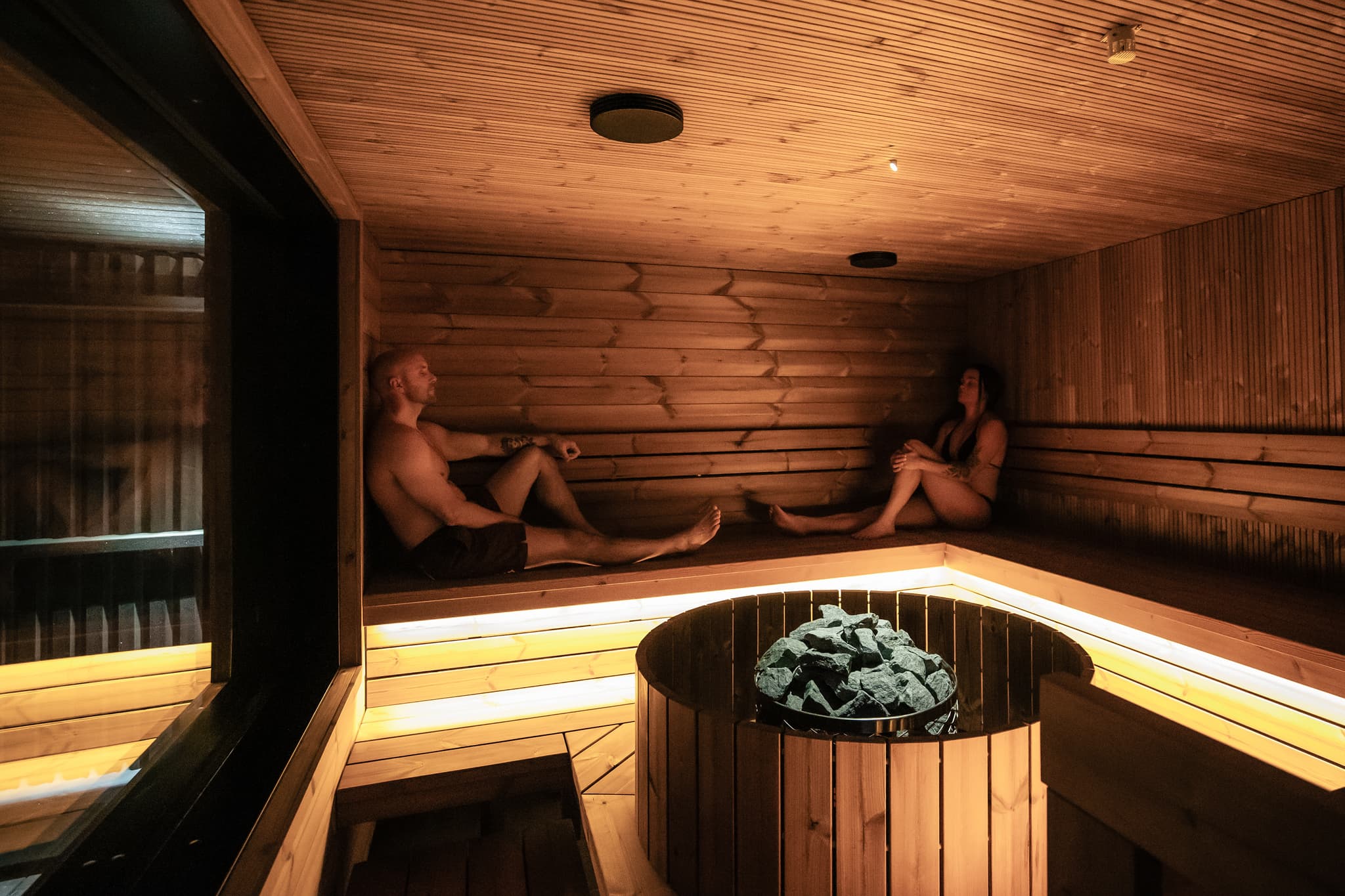 Two people sit inside a warmly lit wooden sauna beside a stone heater.