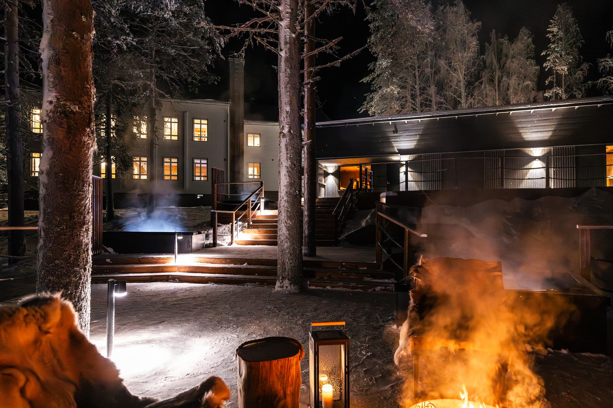 A snowy outdoor spa area glows at night with lit buildings, steam rising from heated pools, and firelight in the foreground among tall trees.