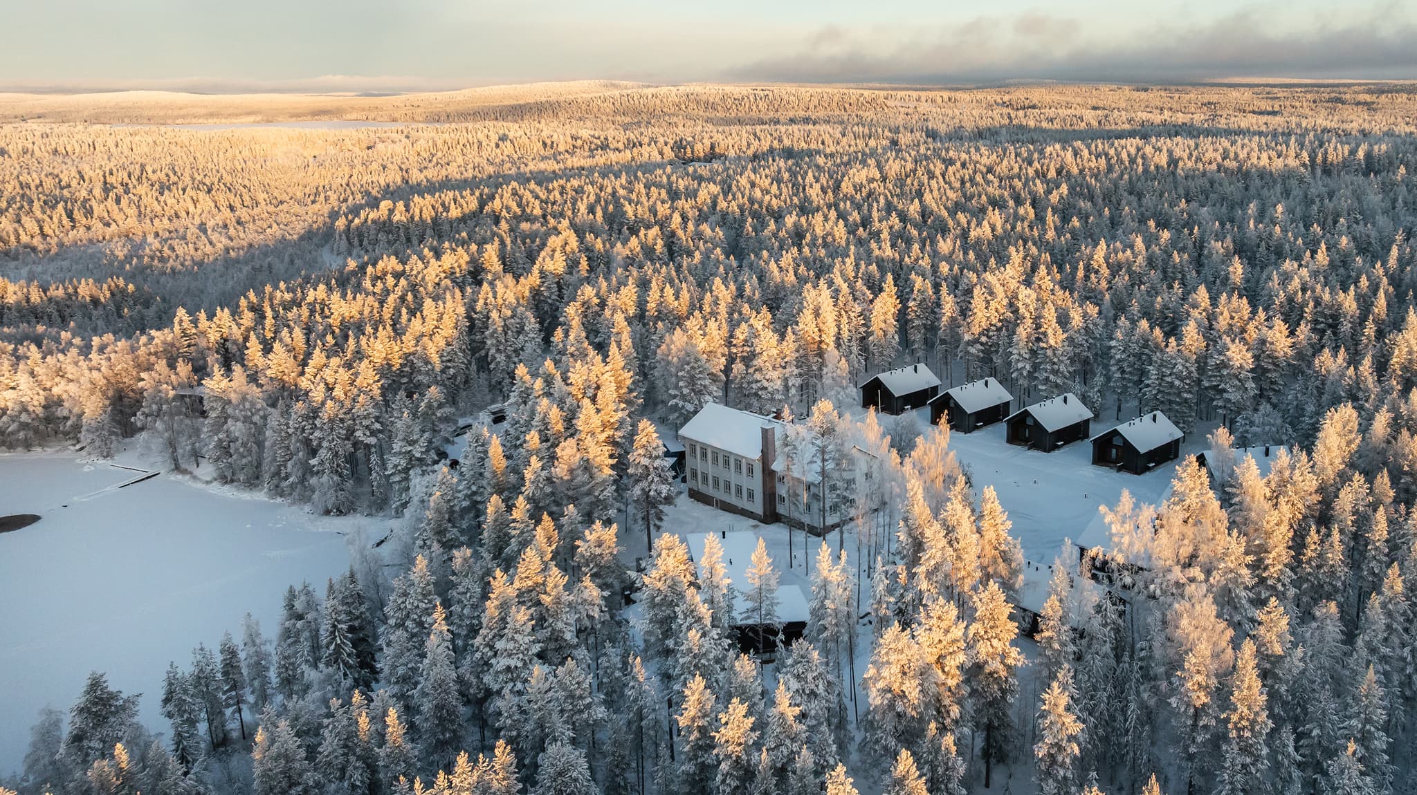 An aerial view shows a snow-covered retreat surrounded by frosted pine forest in winter light.
