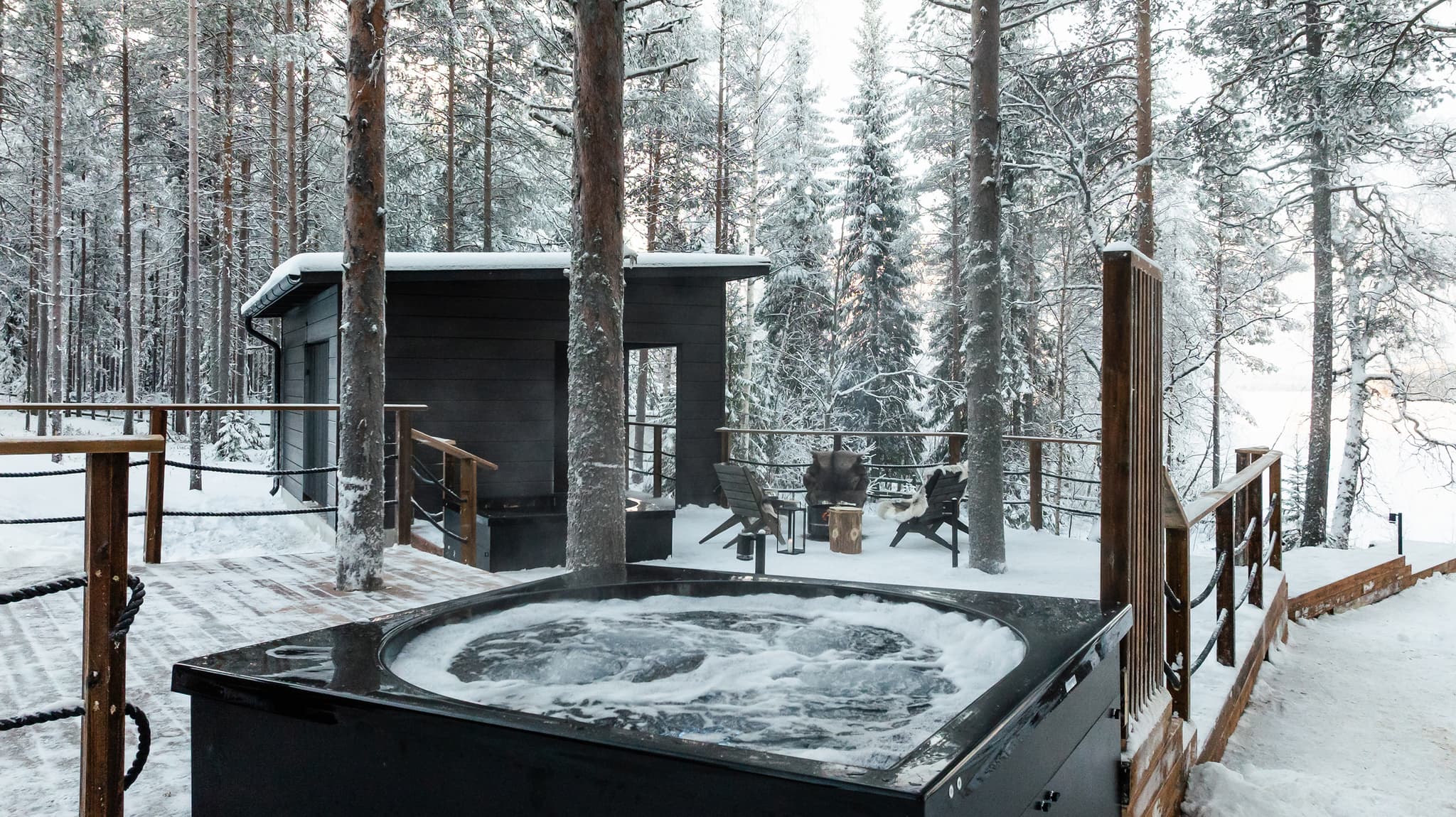 A snow-covered outdoor hot tub sits on a wooden deck in a snowy forest with lounge chairs and a small cabin nearby.