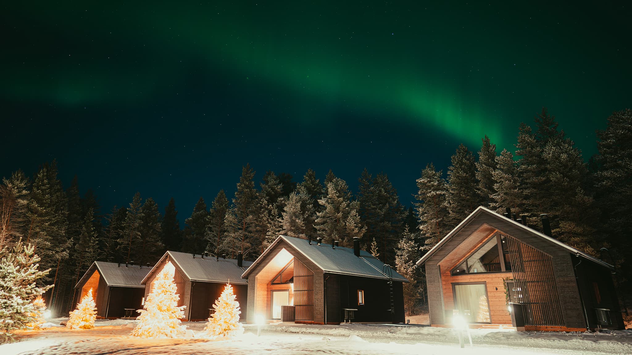A row of snow-covered wooden cabins stands beneath a starry night sky with green aurora above a forest.