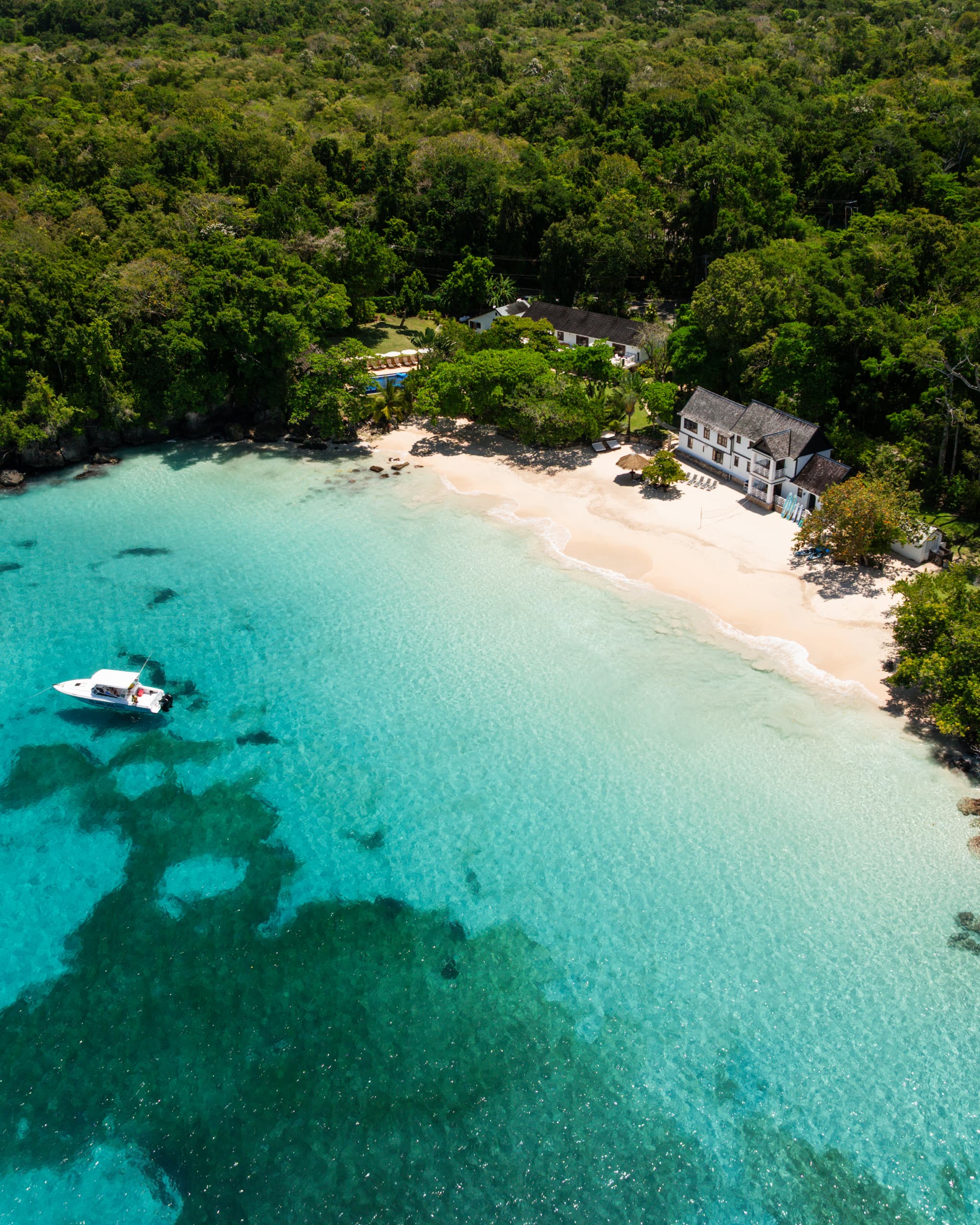 An aerial view shows a small sandy beach with clear turquoise water, a white beachfront villa, and dense green tropical forest behind it, with a small boat anchored offshore.