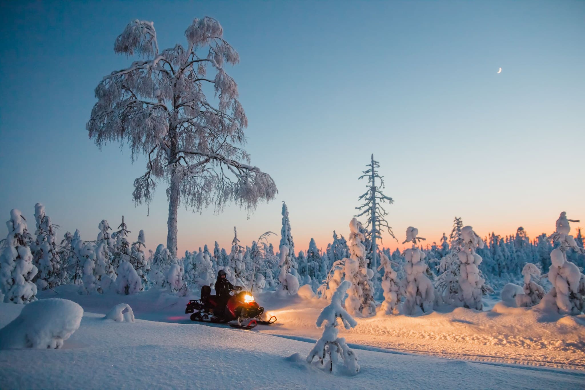 A snowmobiler rides through a snow-covered forest at dusk with the headlights glowing against a blue and orange winter sky.