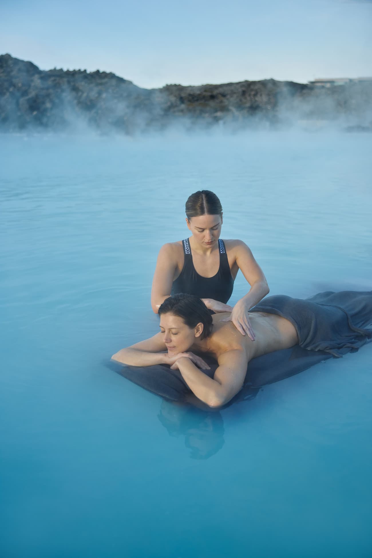 Two people receiving a massage in a calm outdoor pool or hot tub with steam rising and mountains in the background.