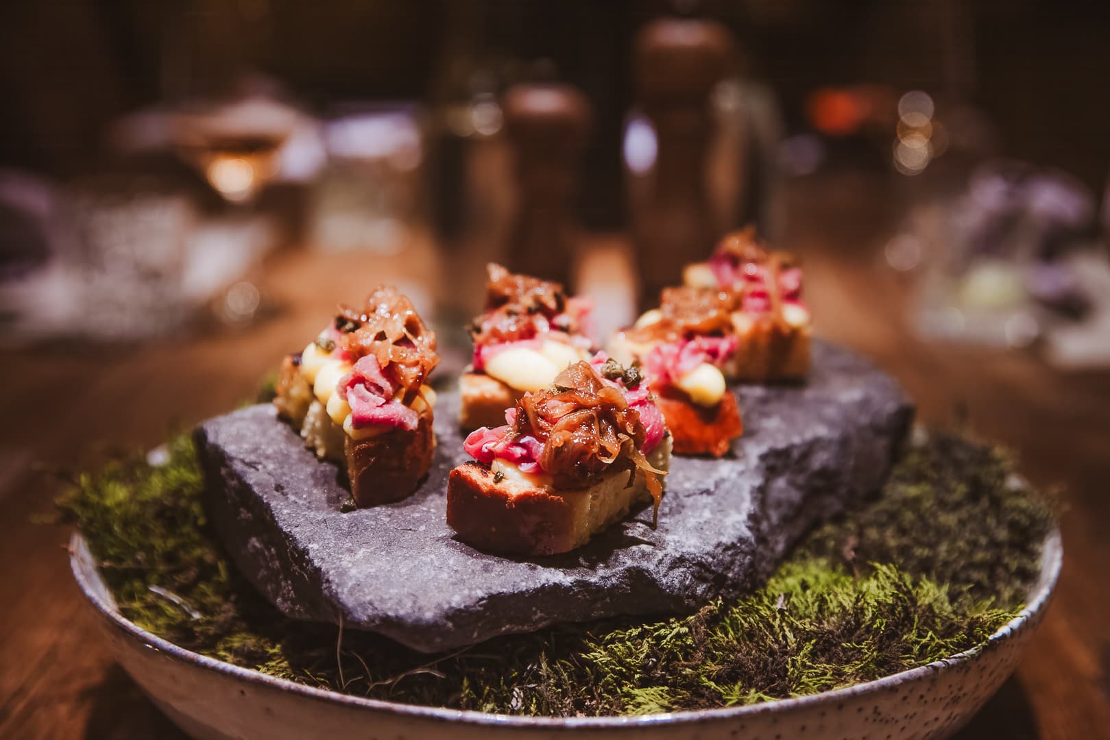 Close-up of bite-sized plated canapés with colorful toppings on a stone slab in a warmly lit setting.