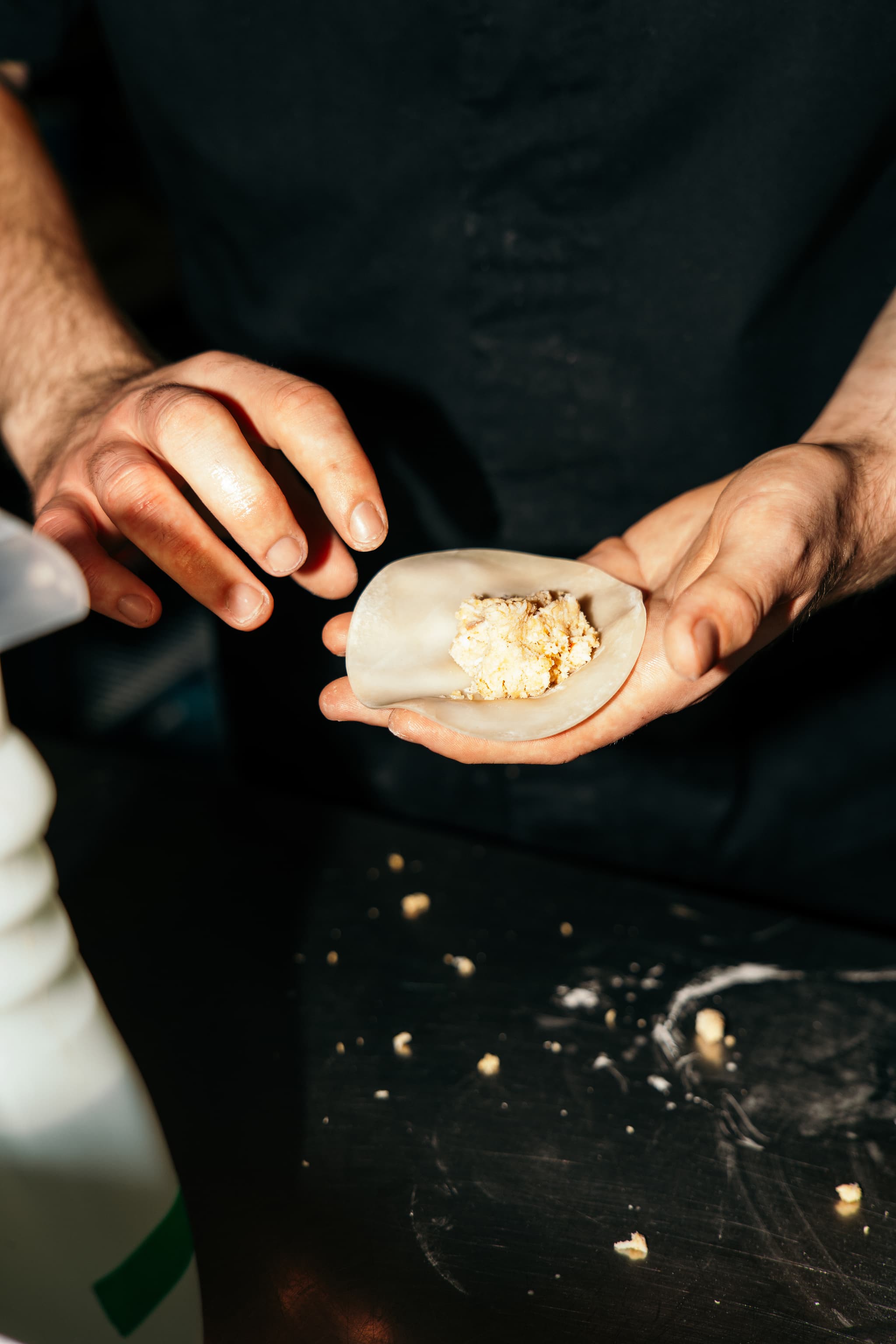 A pair of hands shapes a small dumpling wrapper around a crumbly filling on a dark work surface.