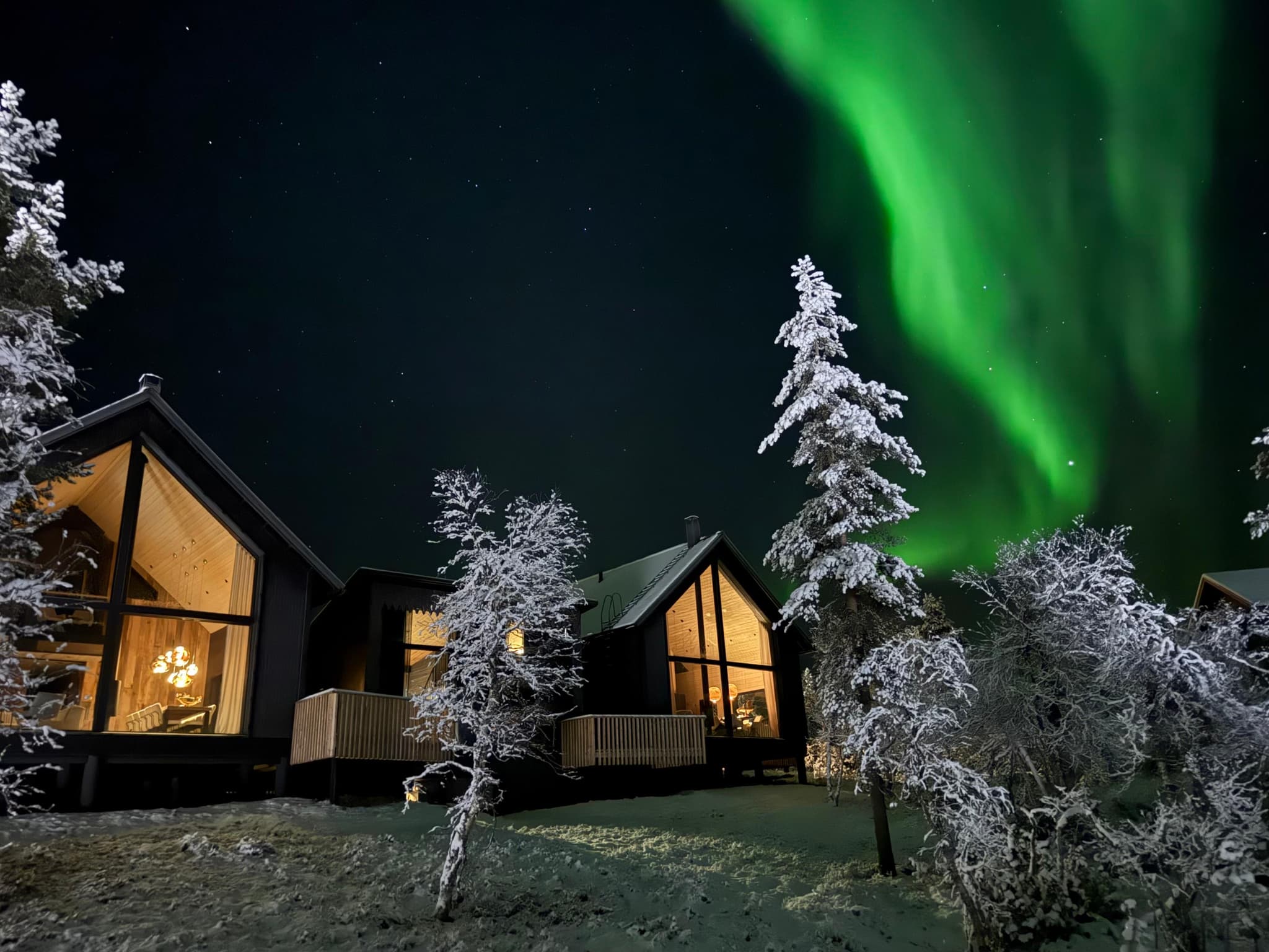 Snow-covered modern cabins glow warmly beneath a vivid green aurora in a dark winter night sky.