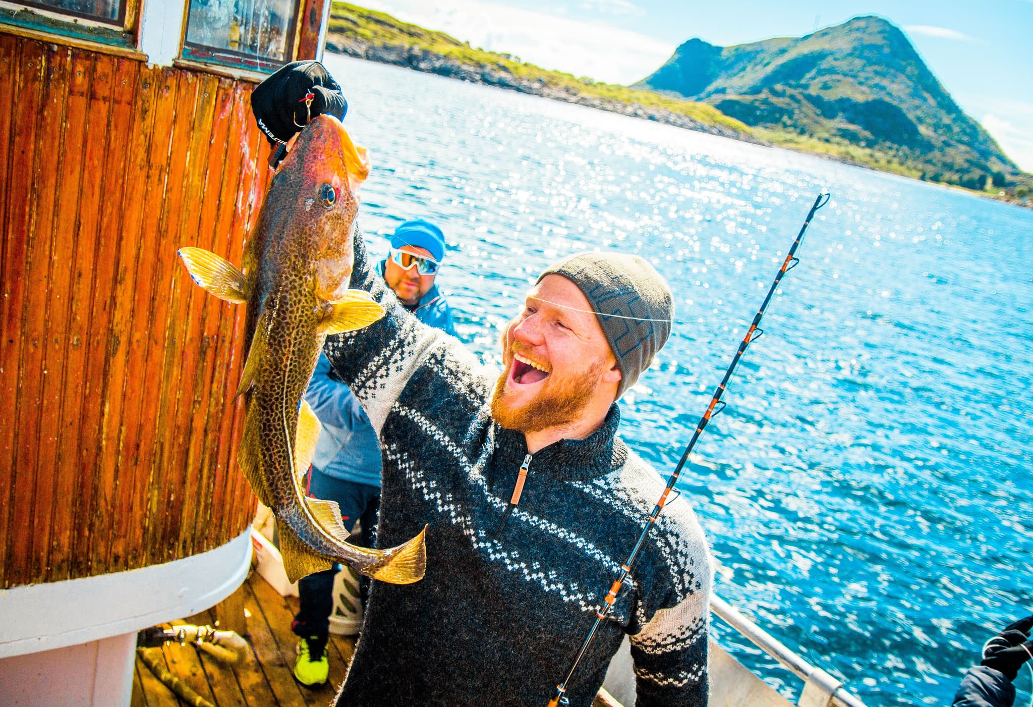 Three people on a boat smile as one holds up a freshly caught fish against a bright blue sea and rugged mountains.