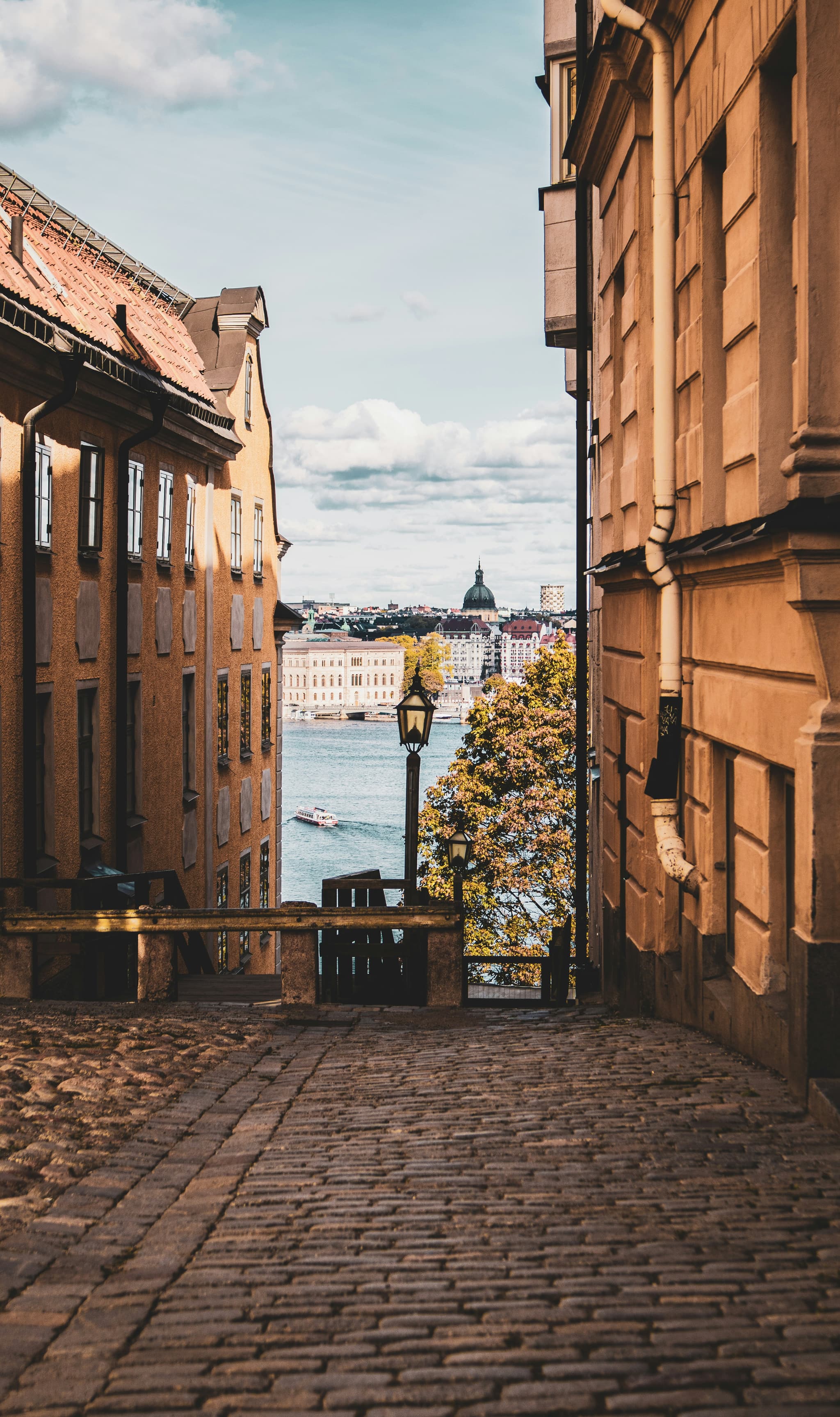 A cobblestone street between old buildings opens to a waterfront view with a distant city skyline and a domed church across the water.