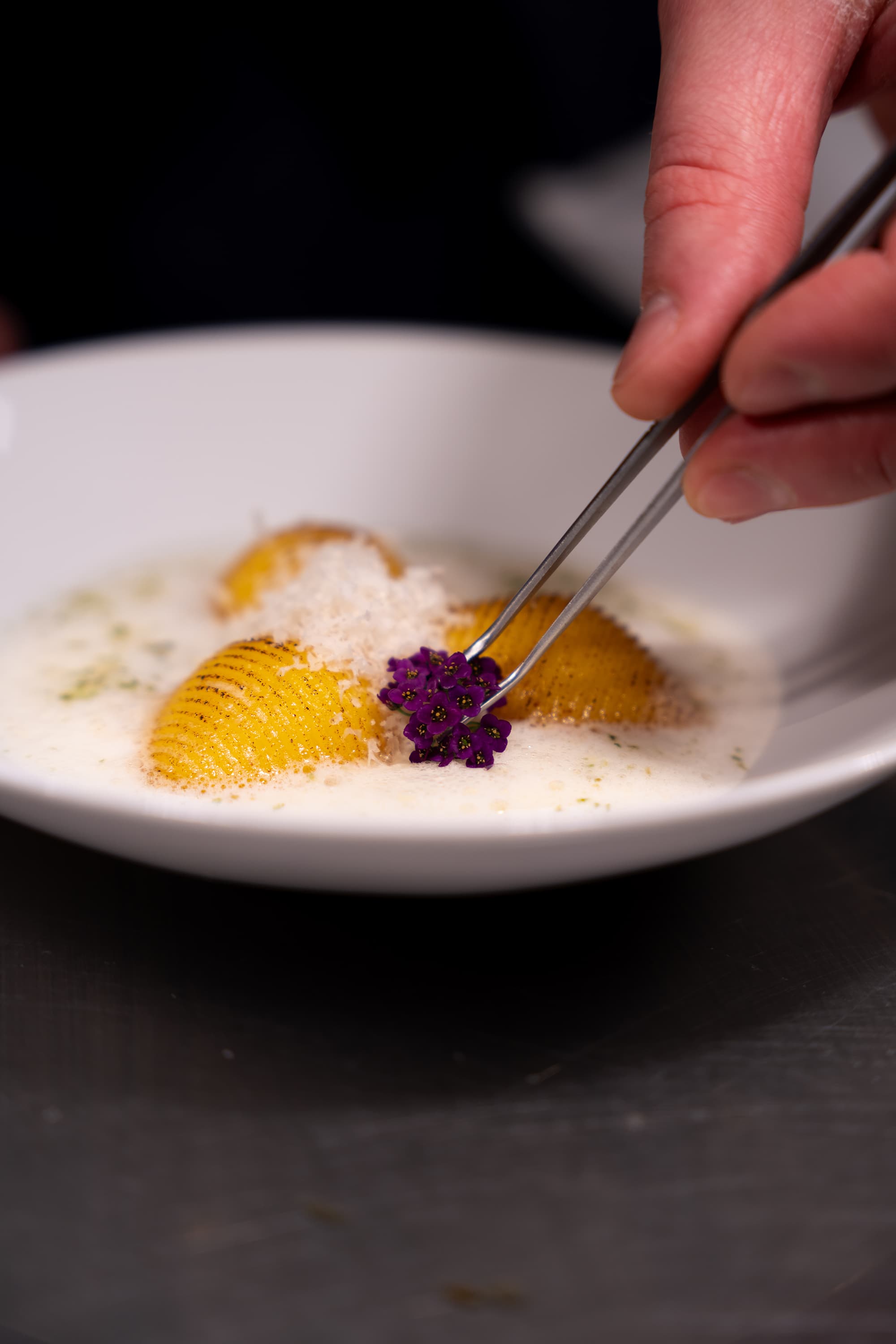A hand uses tweezers to place a small purple flower garnish onto a plated fine-dining dessert in a white bowl.