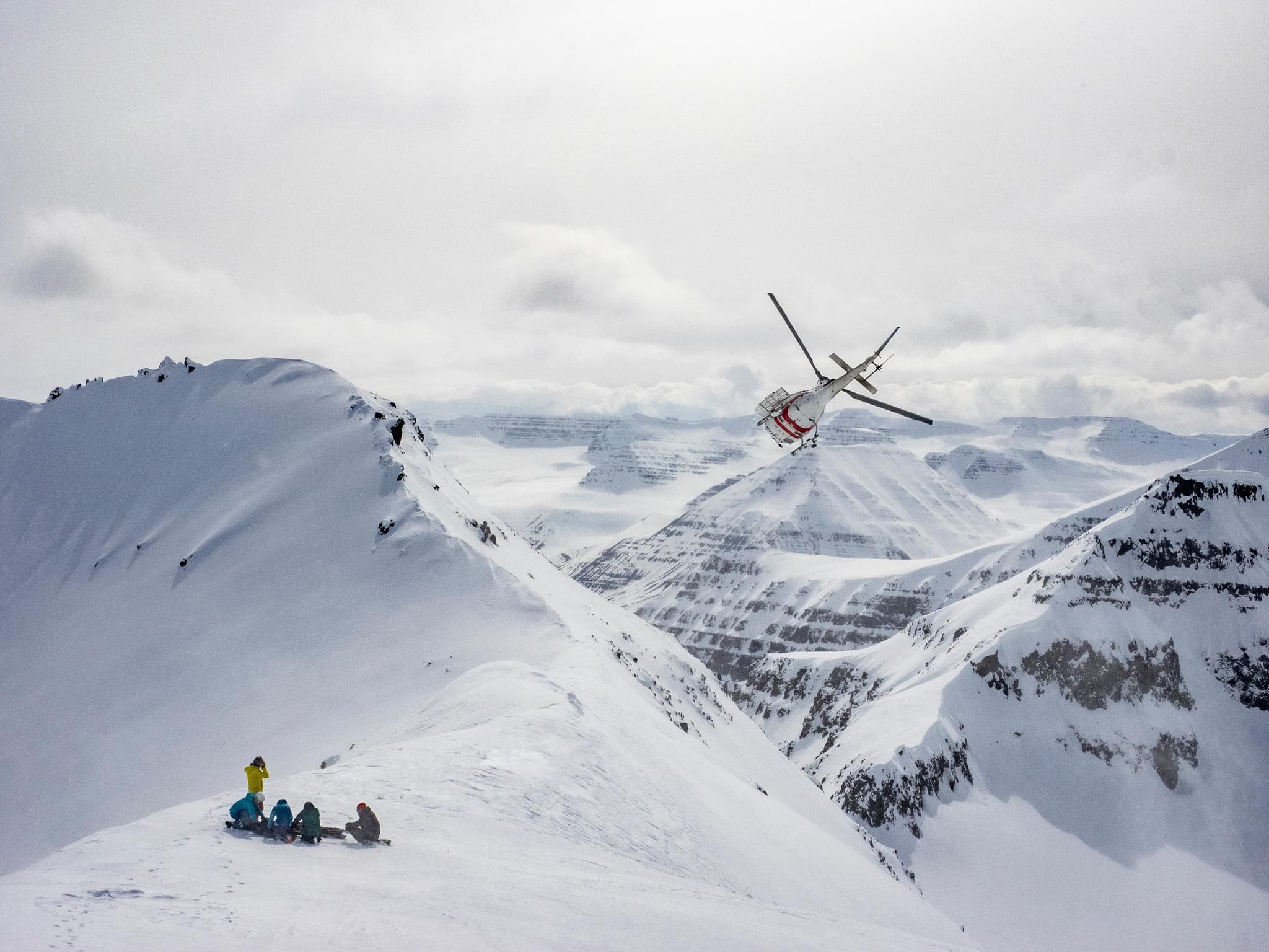 A helicopter flies over a snowy mountain ridge while a small group of people stands on the foreground slope.