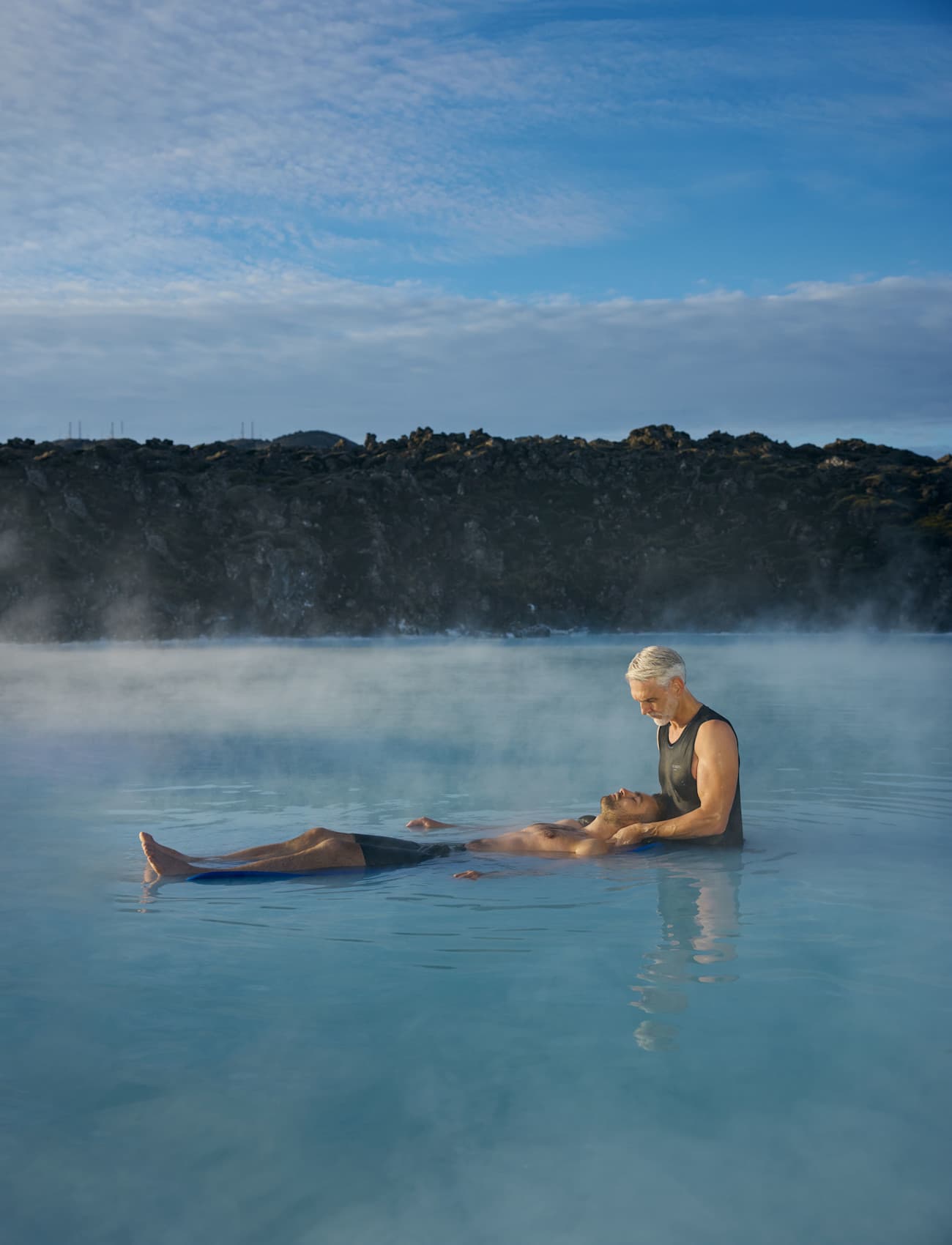 Two people receiving and giving an in-water massage in a calm outdoor pool or hot spring, with mist rising and hills in the background under a blue sky.