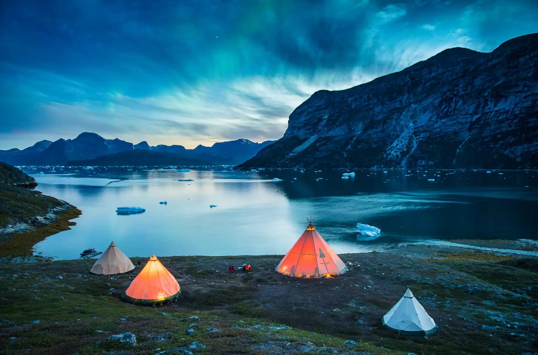 A remote lakeside campsite with glowing tents set beside calm water, floating ice, and steep mountains under a dramatic blue sky at dusk.