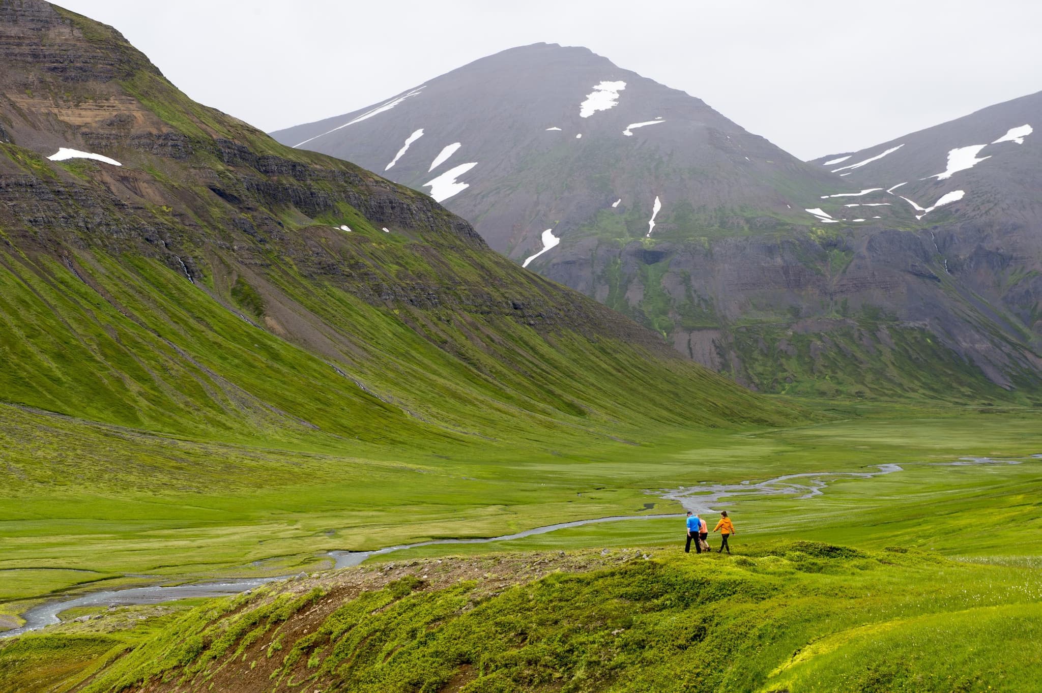 A small group of hikers walks across a bright green valley beside a winding stream, with steep mountains and patches of snow rising in the background.