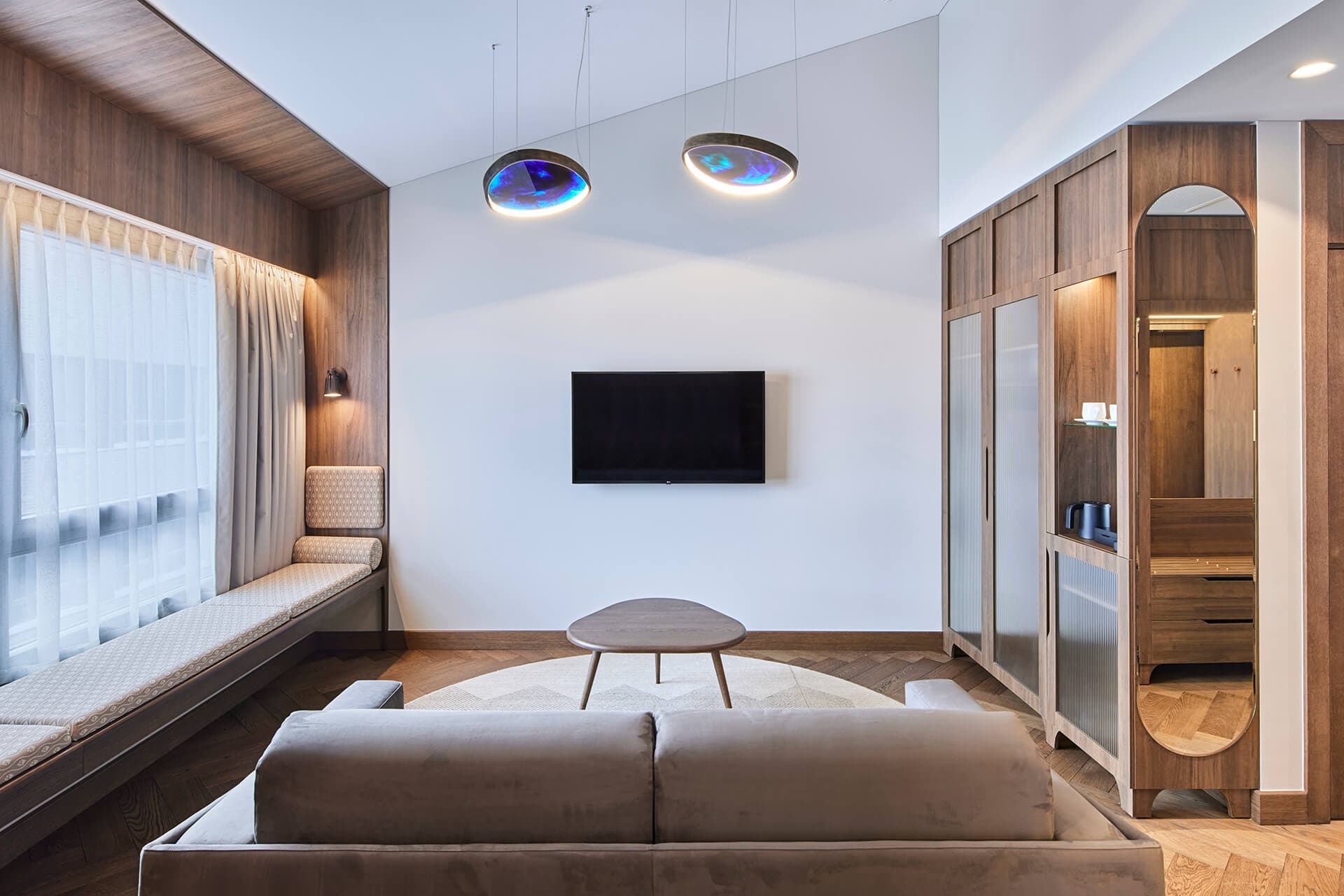 A modern living room with a sofa, window seat, wall-mounted TV, and wood-paneled cabinetry beneath pendant lights.