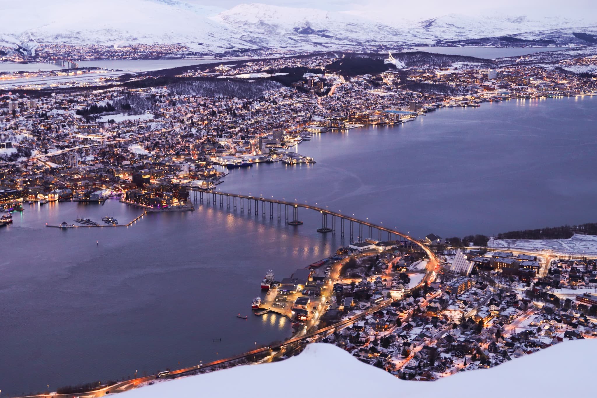 An aerial winter view shows a snowy coastal city with a long bridge crossing the water and mountains in the background at dusk.