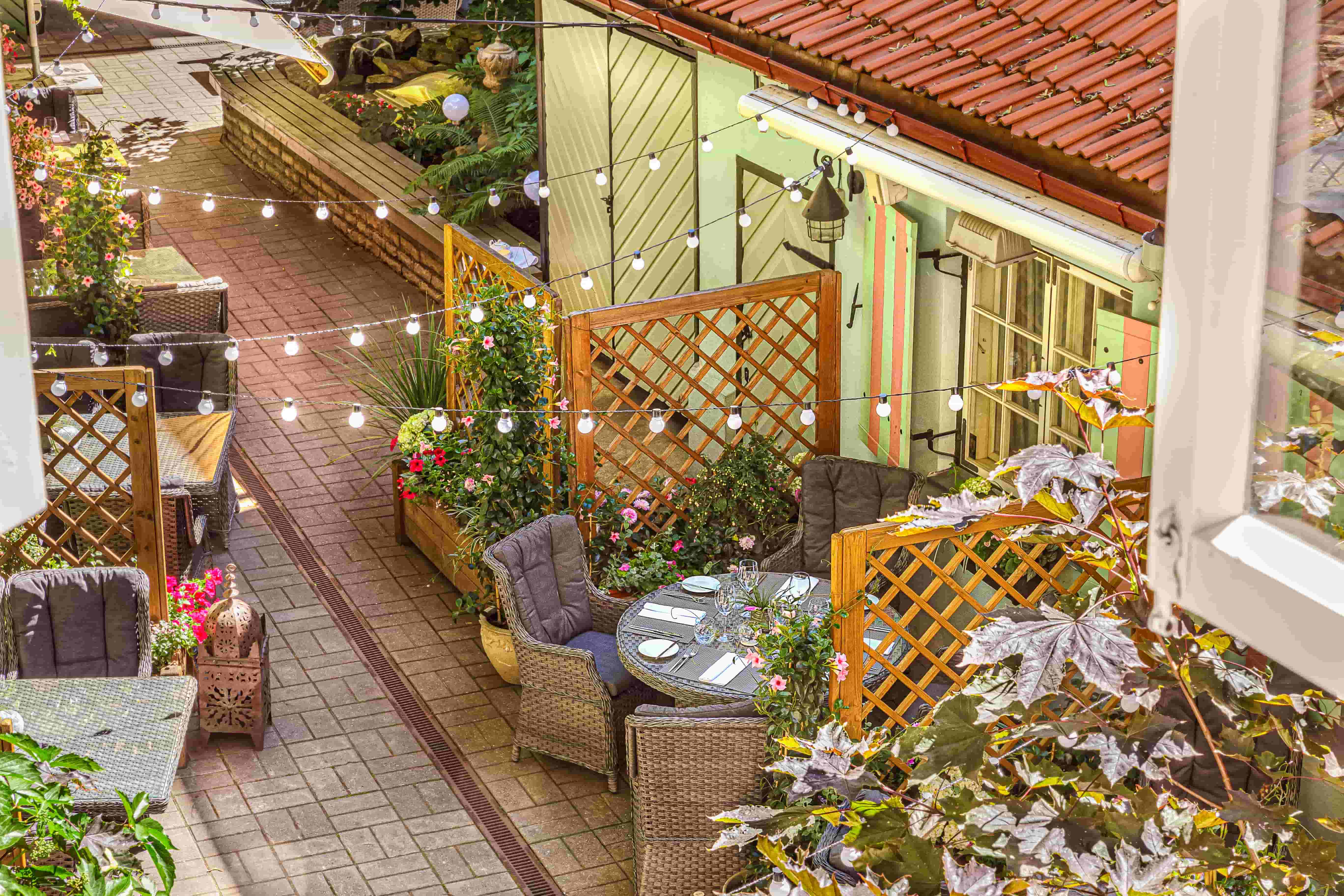 An outdoor courtyard with wicker dining chairs, small tables, potted flowers, string lights, and a pastel building with a red tiled roof.
