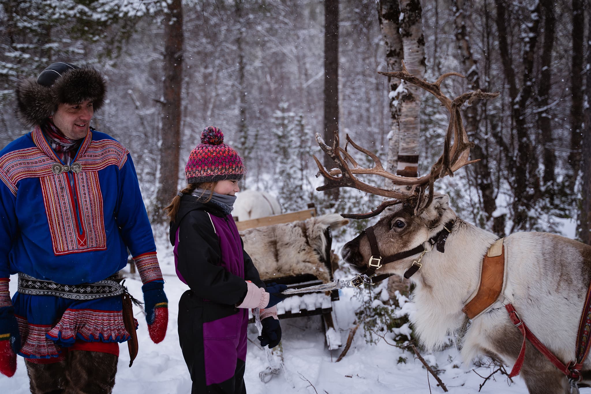A man in traditional dress and a child stand beside a reindeer in a snowy forest.