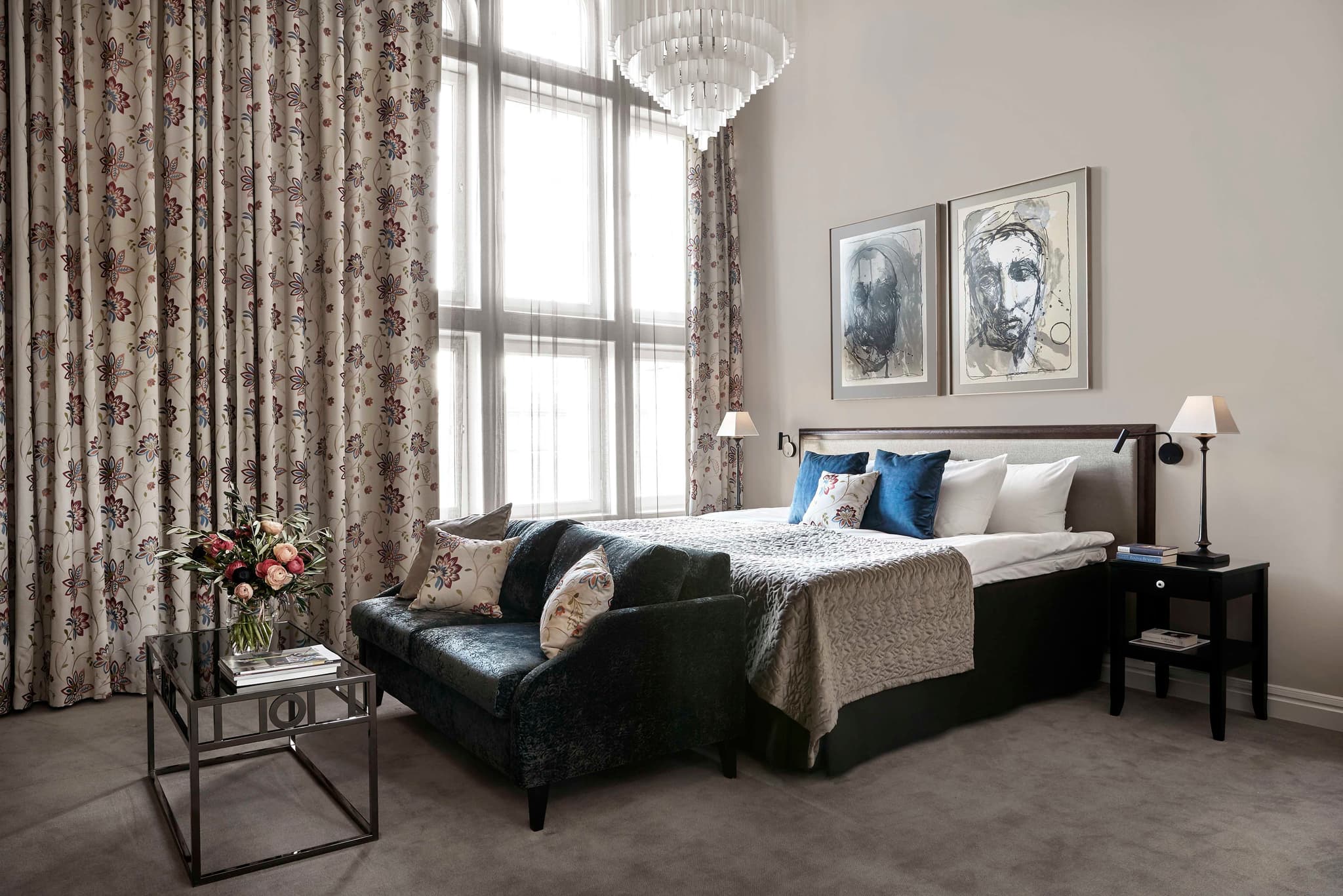 A bright, elegantly styled bedroom features a bed, patterned curtains, a sofa at the foot of the bed, and framed artwork on a pale wall.