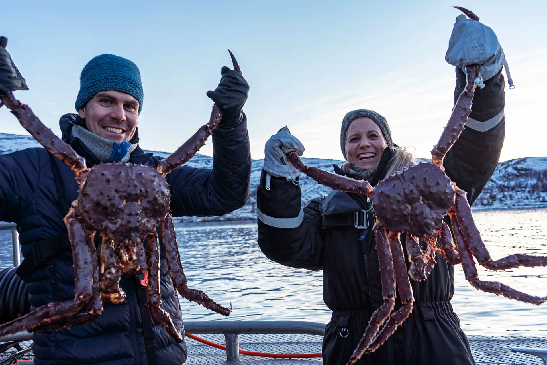 Two bundled-up people on a boat hold up large king crabs against a snowy coastal backdrop.