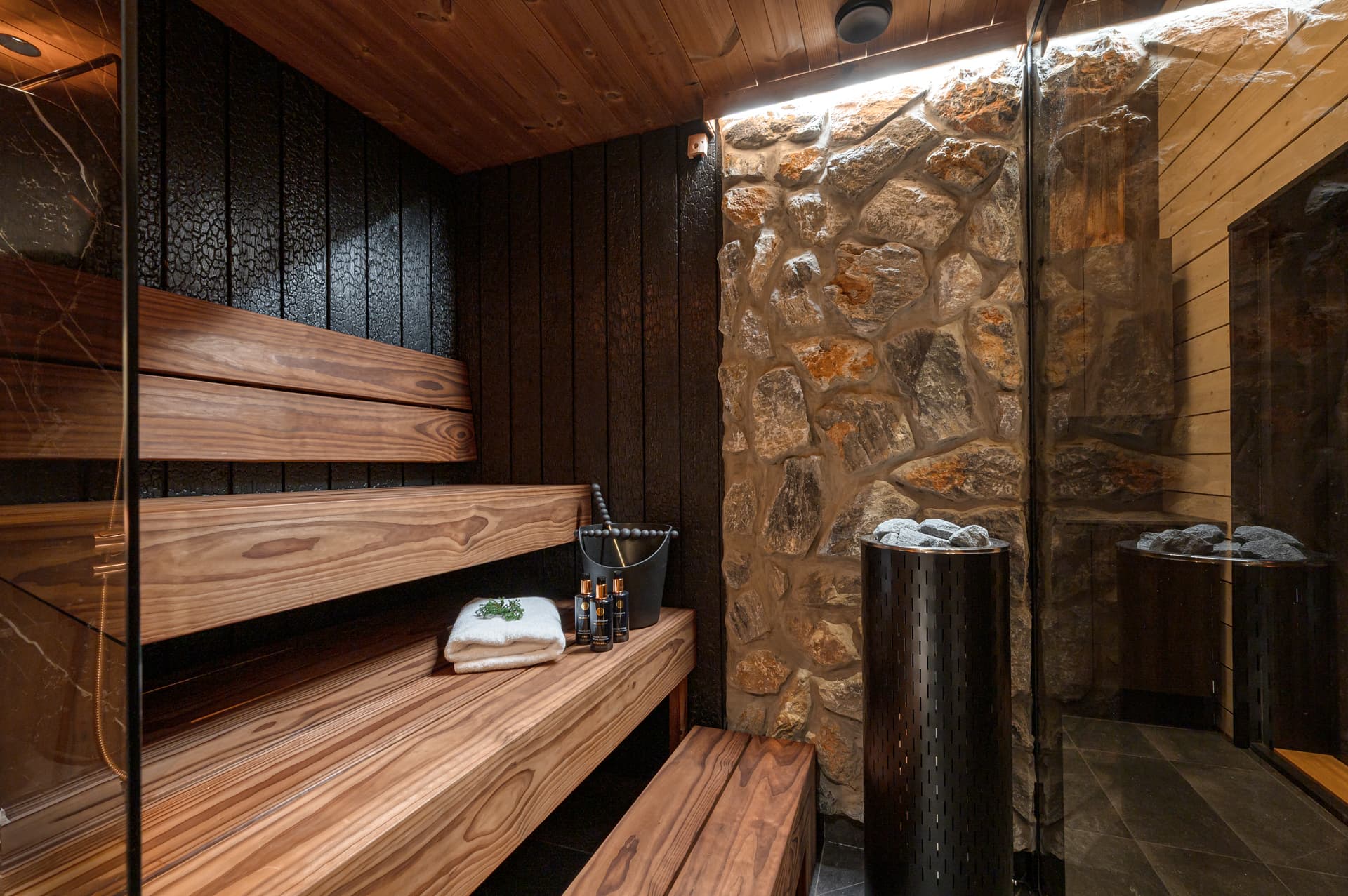 A rustic wooden sauna interior features bench seating, stone walls, and sauna accessories beside the heater.