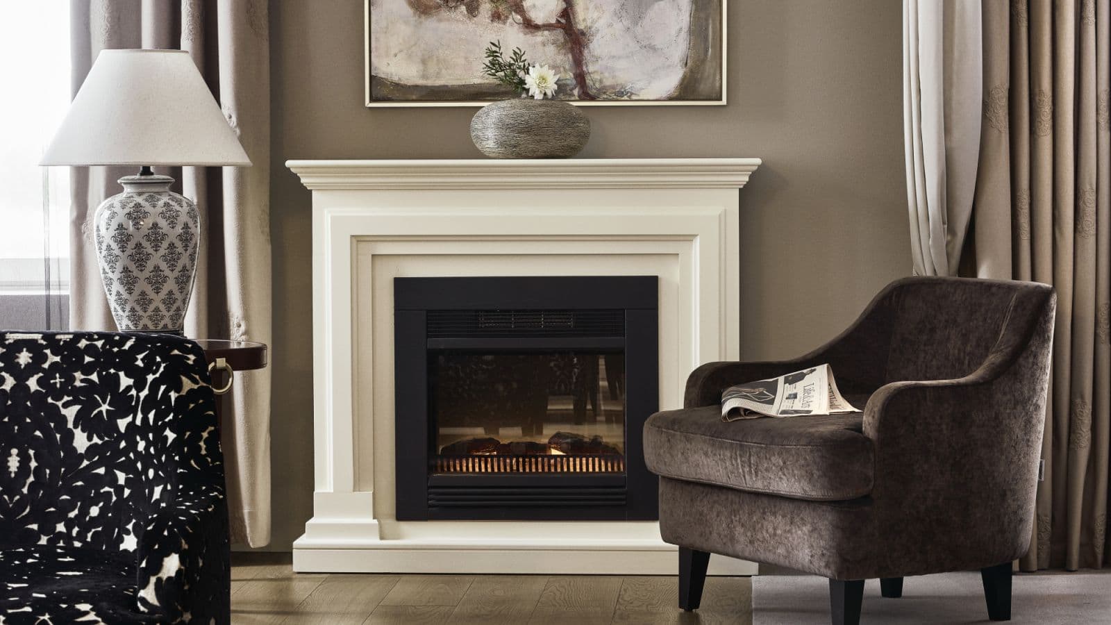 A stylish living room features a white fireplace, two upholstered armchairs, a patterned table lamp, and layered curtains in neutral tones.