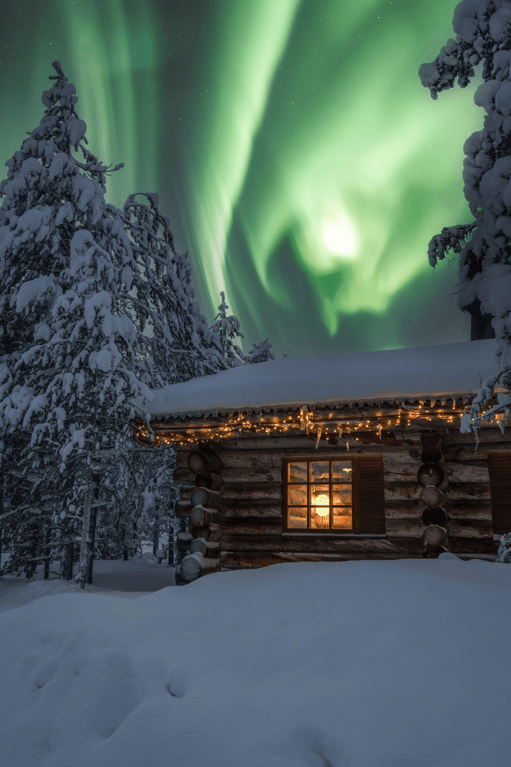 Snow-covered pine trees frame a log cabin glowing with warm lights beneath a vivid green aurora in the night sky.
