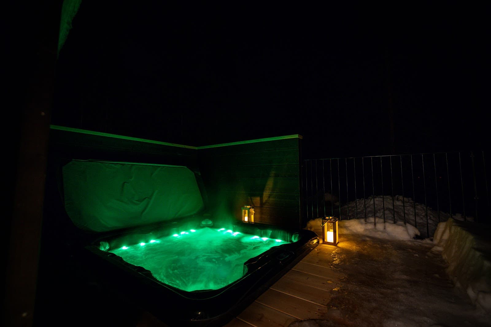 Night exterior with a green-lit hot tub/spa in a snowy courtyard beside lantern lights and a dark fence wall.