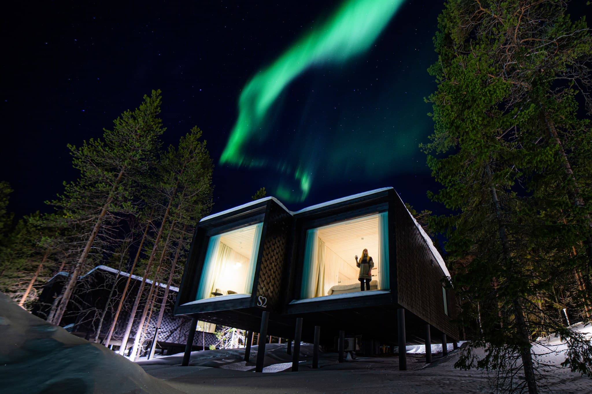 A modern treehouse cabin stands among snowy pine trees beneath a vivid green aurora in a starry night sky, with a person visible inside one lit window.