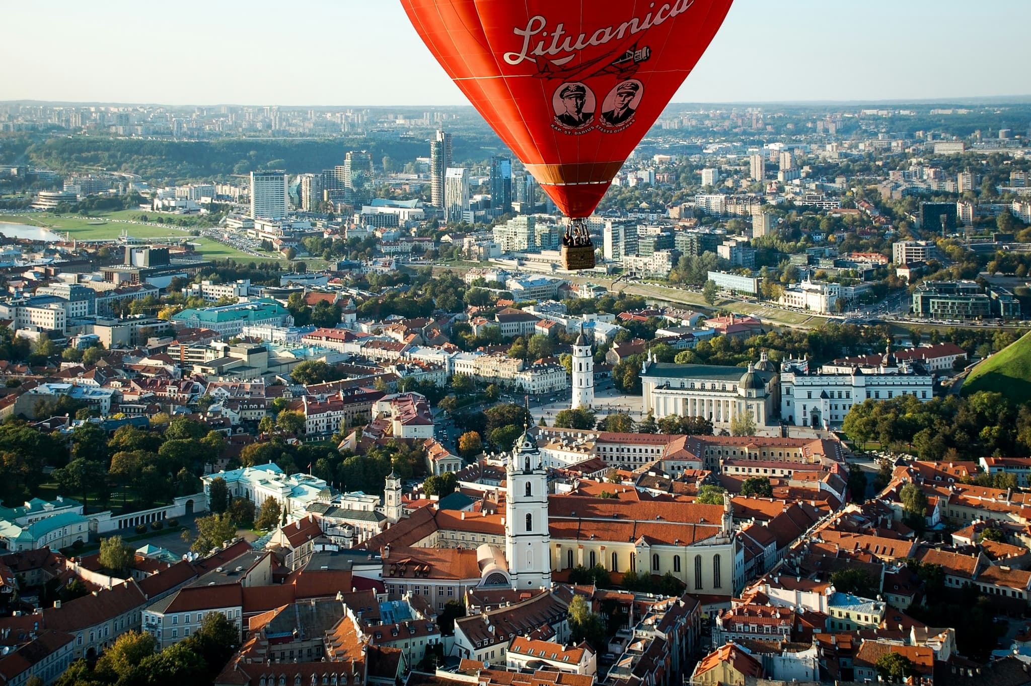 A red hot air balloon floats above a dense historic cityscape with red-roofed buildings and church towers on a clear day.