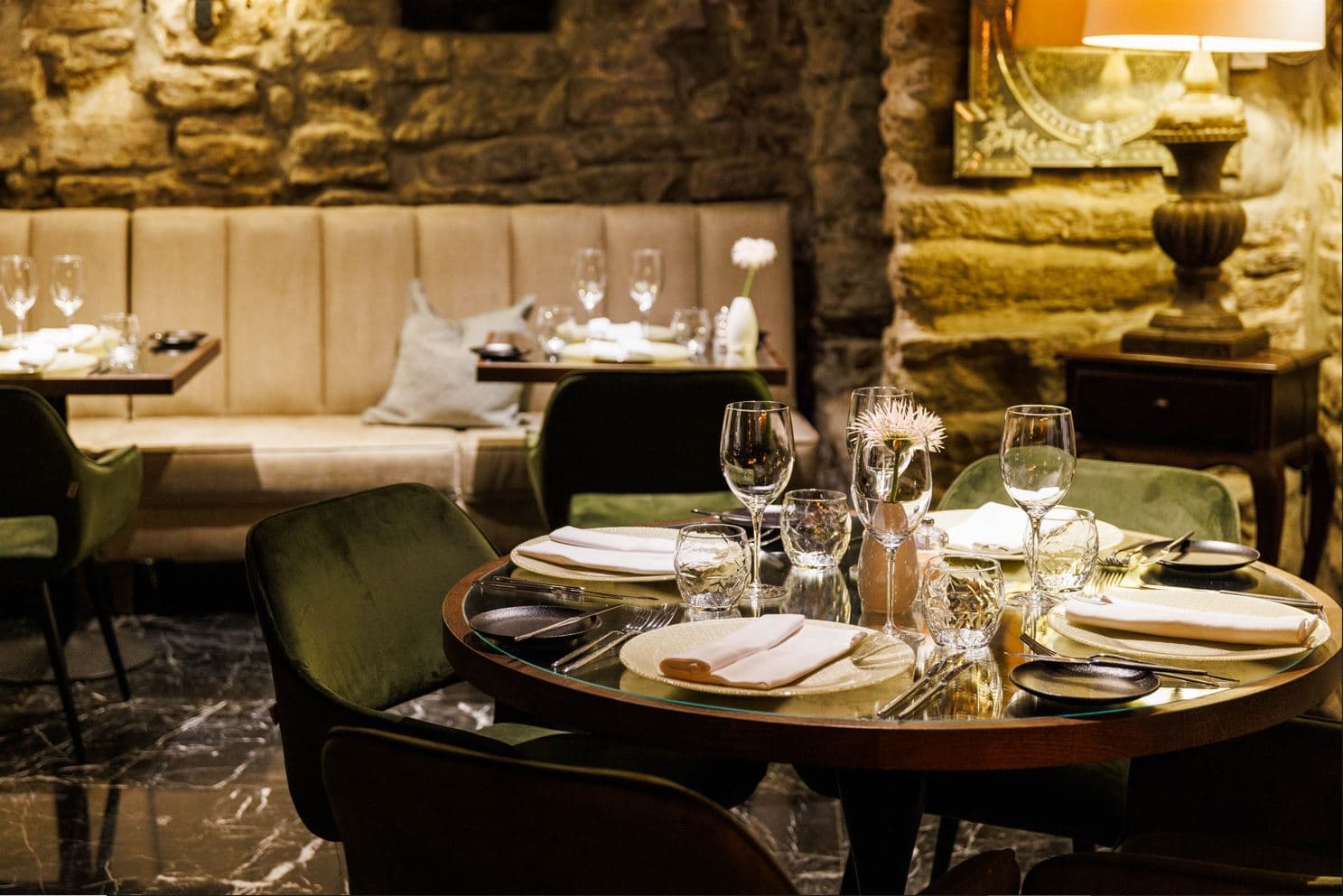 A warmly lit restaurant dining room features set tables with glassware and napkins, upholstered green chairs, and a stone wall backdrop.