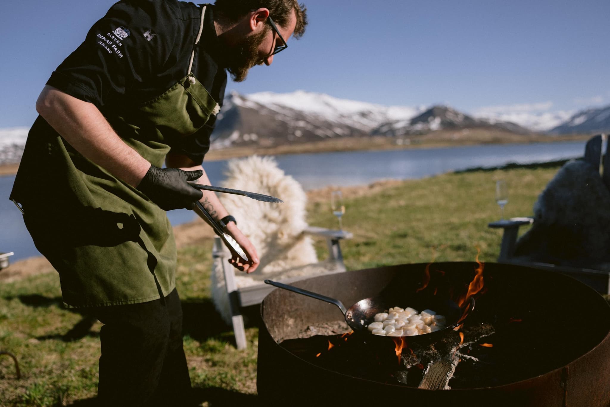 A man in an apron cooks food in a pan over an open fire beside a lake with snow-covered mountains in the background.
