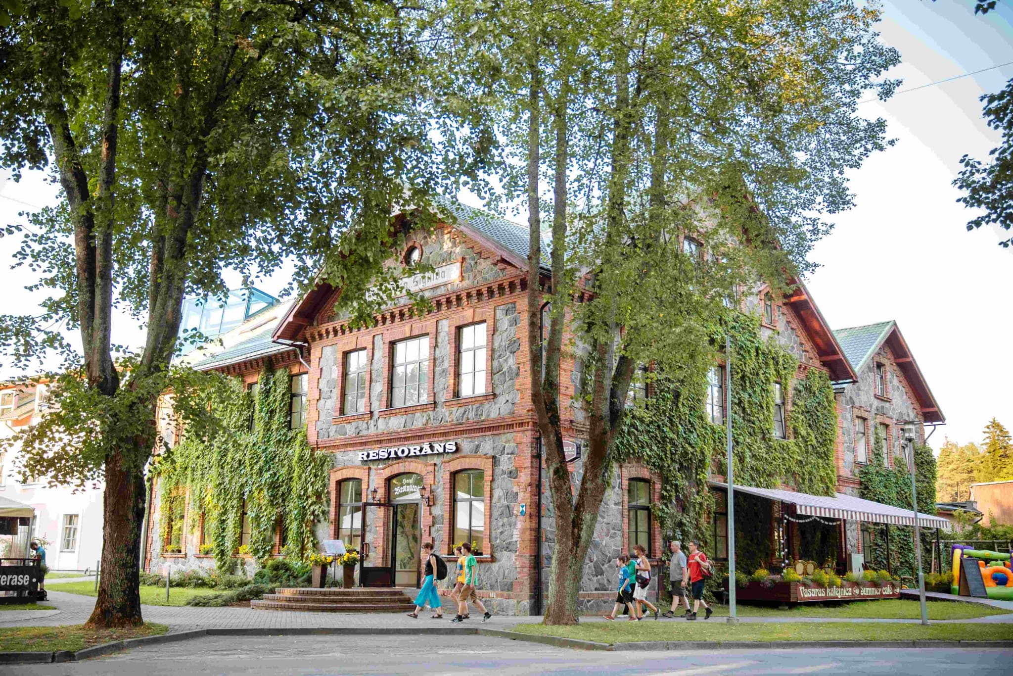 A ivy-covered brick building with a restaurant entrance sits among tall trees while several people walk past on a sunny day.