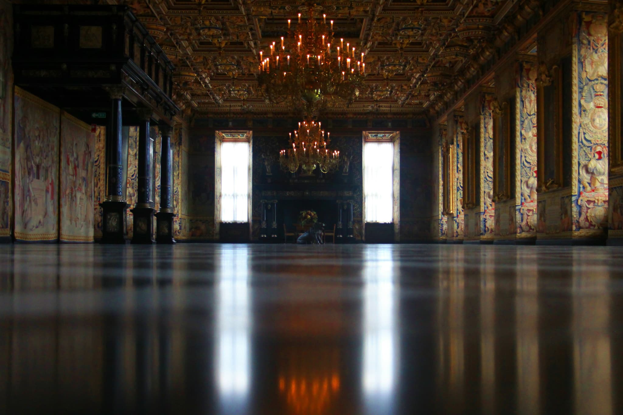 An ornate castle interior with painted walls, chandeliers, tall windows, and a polished floor reflecting the light.