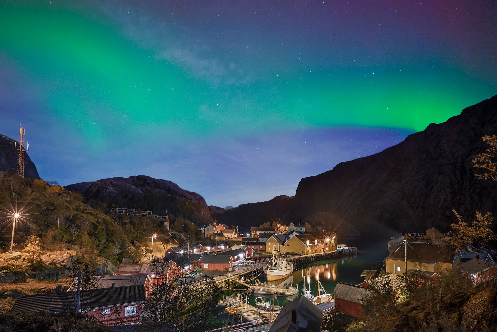 A green aurora borealis glows over a small harbor village nestled between dark mountains at night.